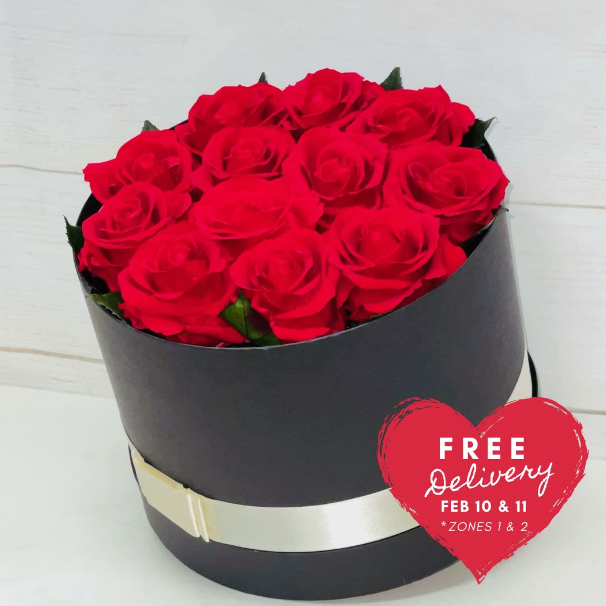 Round black hat box filled with tightly packed red roses and a cream ribbon accent.