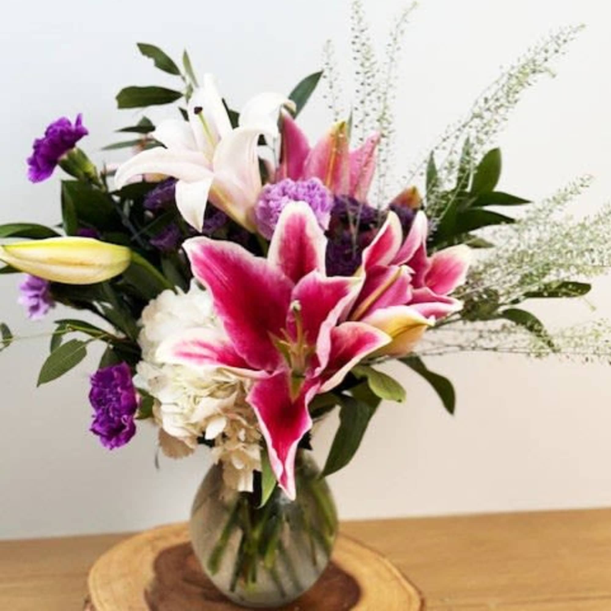 Pink lilies, white blooms, and purple flowers in a clear glass vase on a wooden slice.