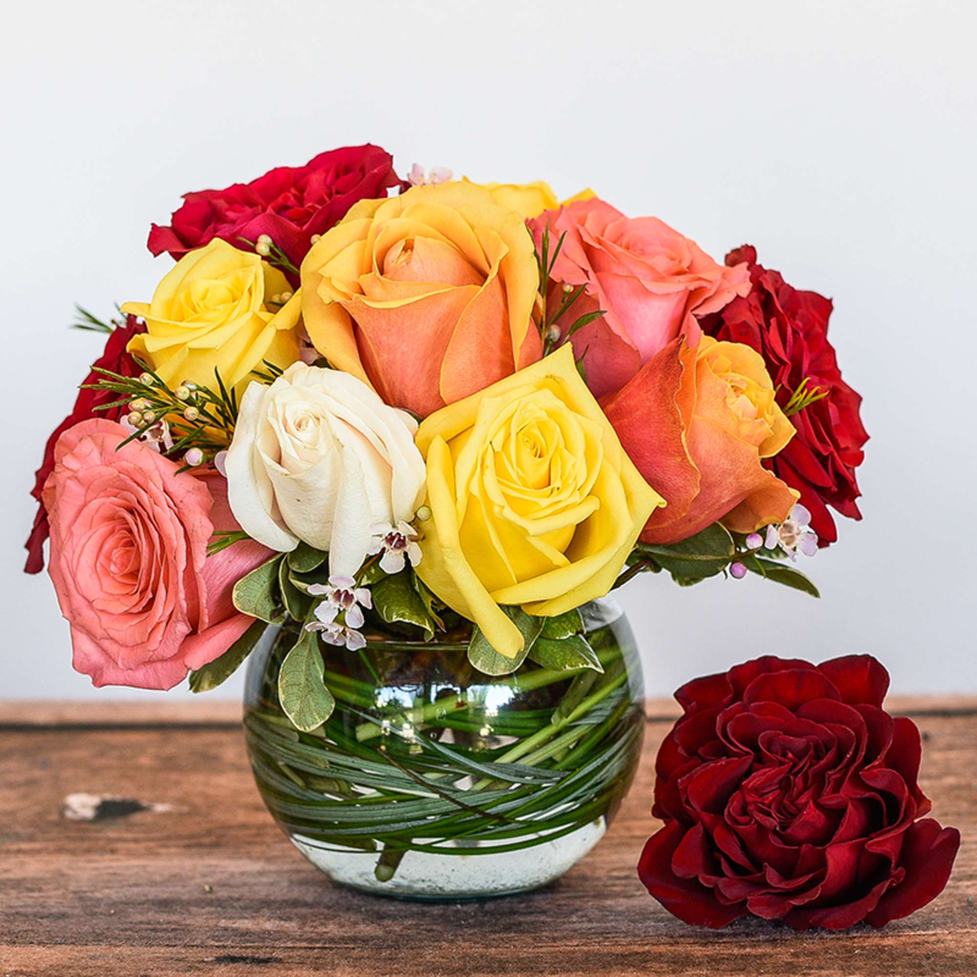 Round glass vase of mixed yellow, red, peach, and white roses with an extra red rose beside it