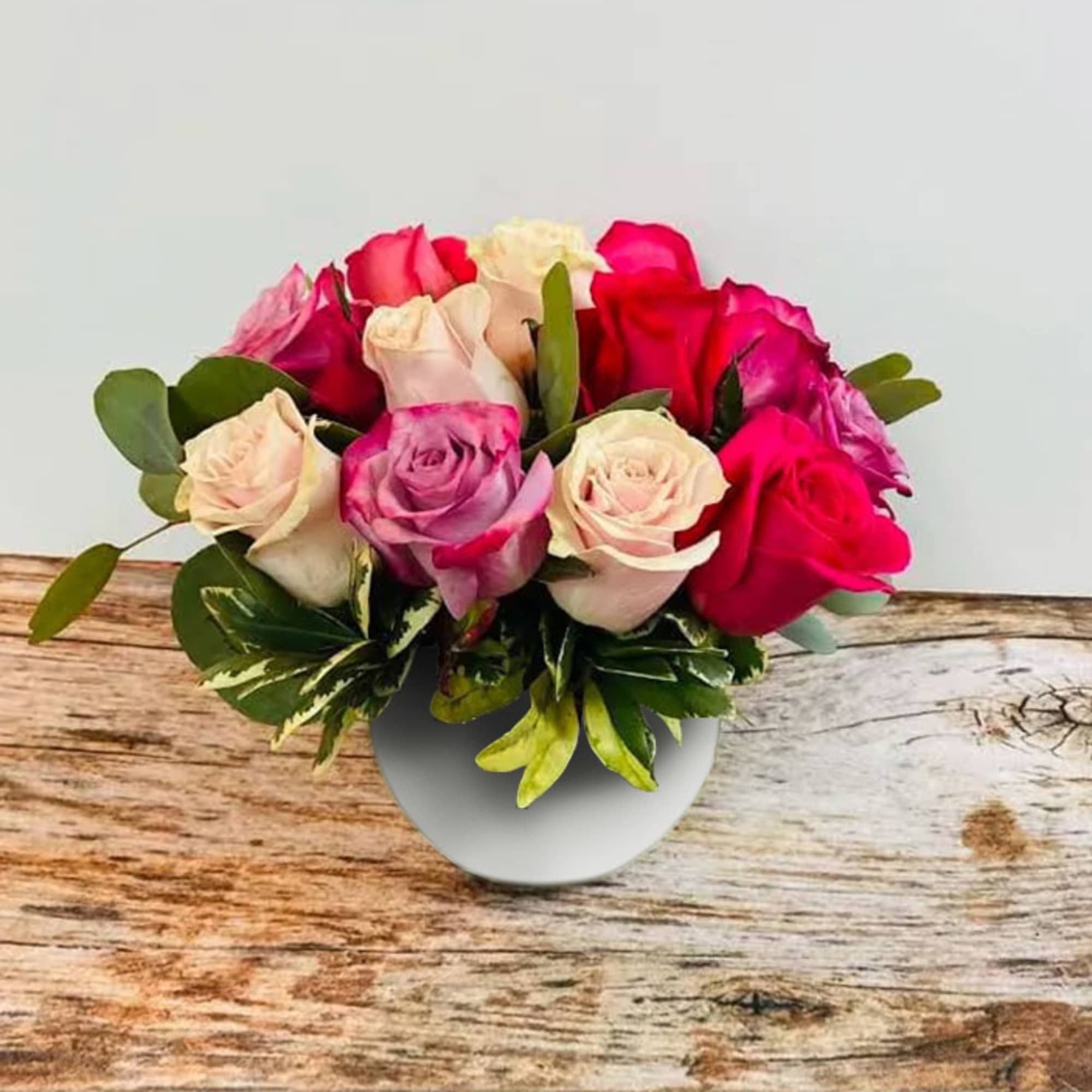 Low round arrangement of pink, red, and cream roses in a white vase on a wooden surface