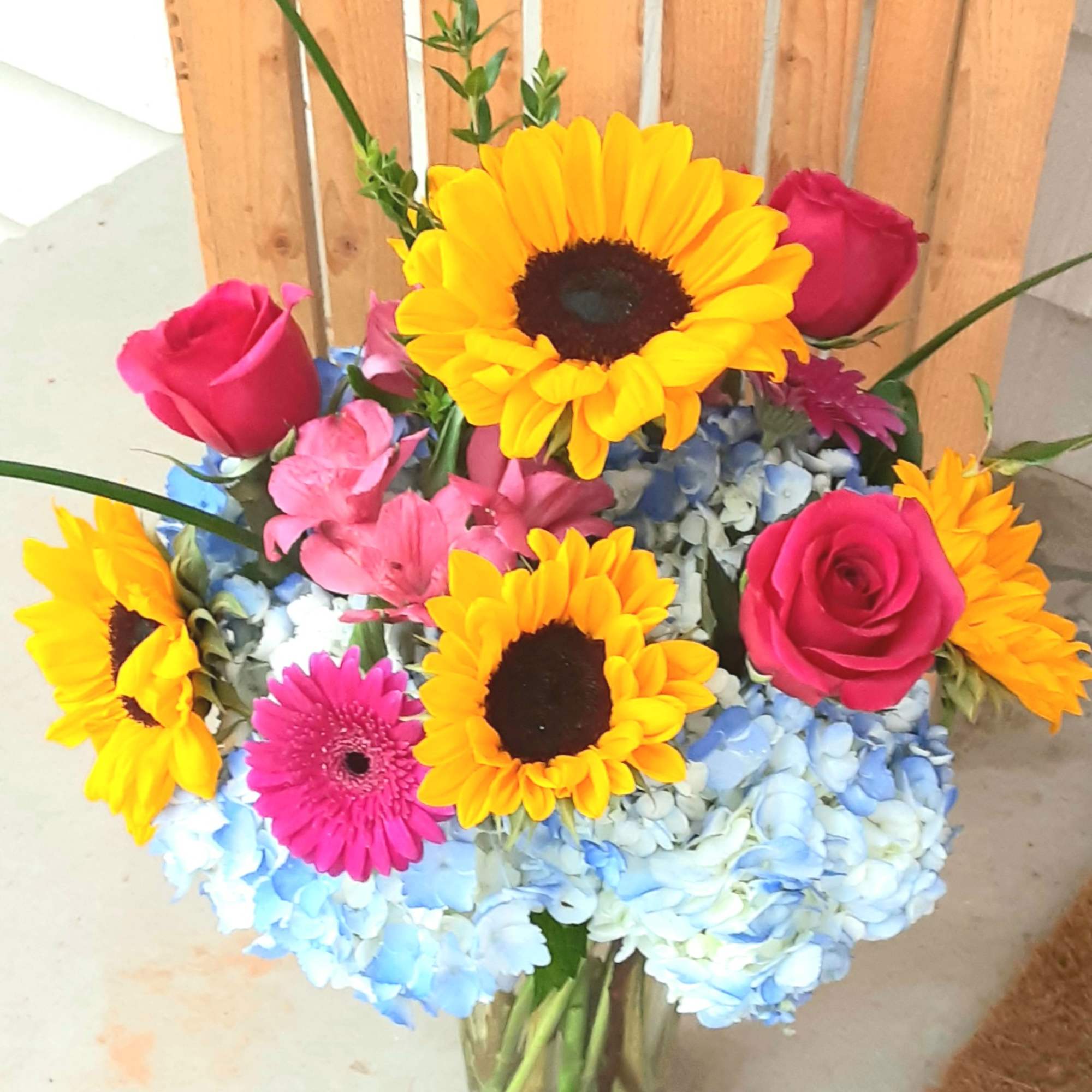 Mixed bouquet with sunflowers, pink roses, blue hydrangeas, and a gerbera daisy in a glass vase