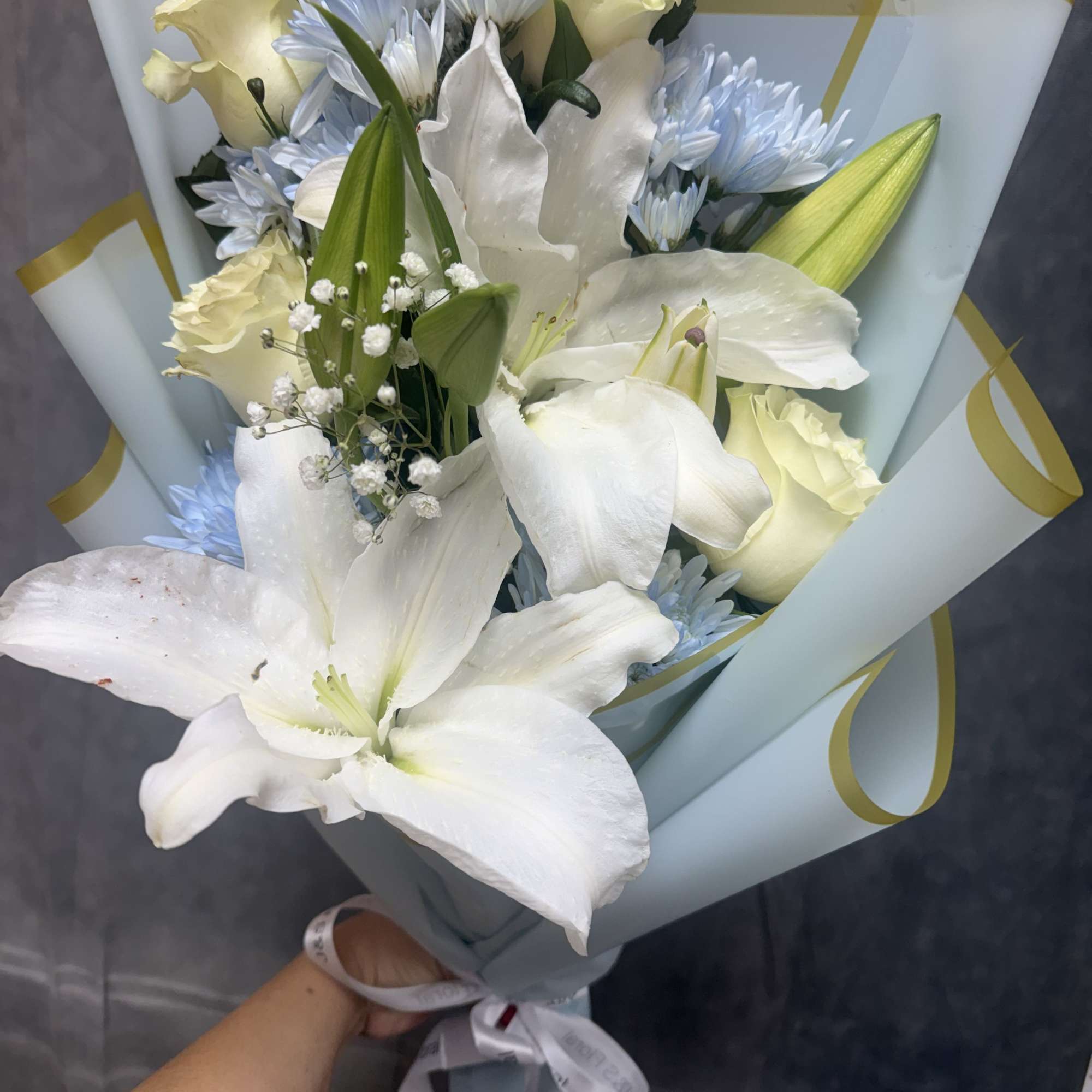 Bouquet of white lilies, pale yellow roses, and blue daisies wrapped in pastel paper