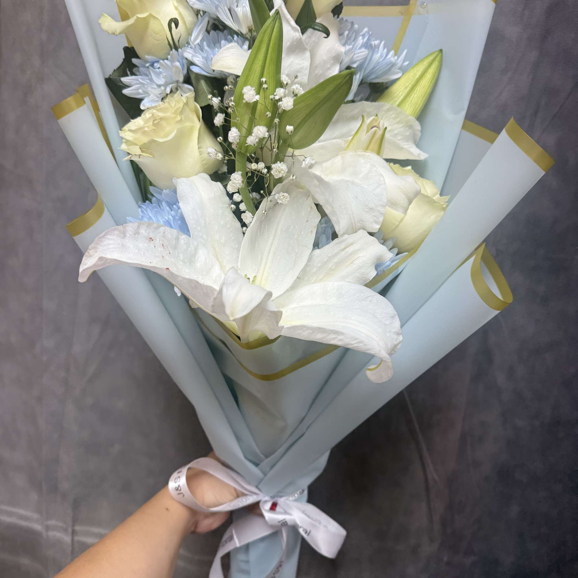 Bouquet of white lilies, pale yellow roses, and light blue flowers wrapped in blue paper