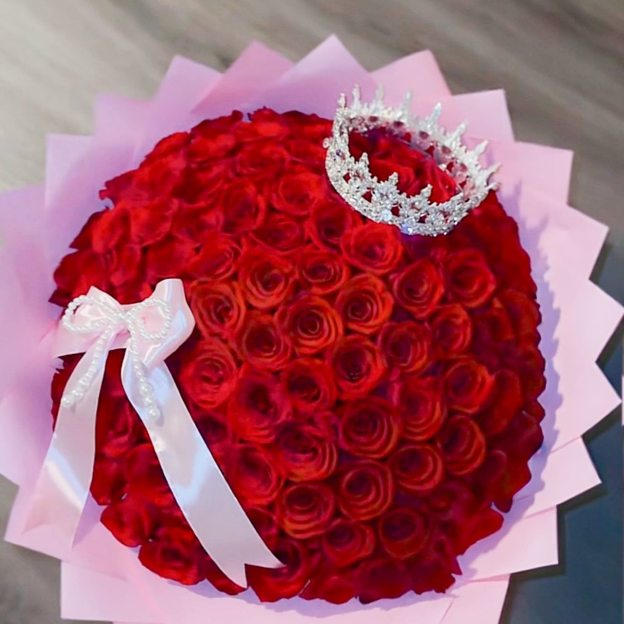 Round bouquet of red roses wrapped in pink paper with a jeweled crown and white ribbon bow