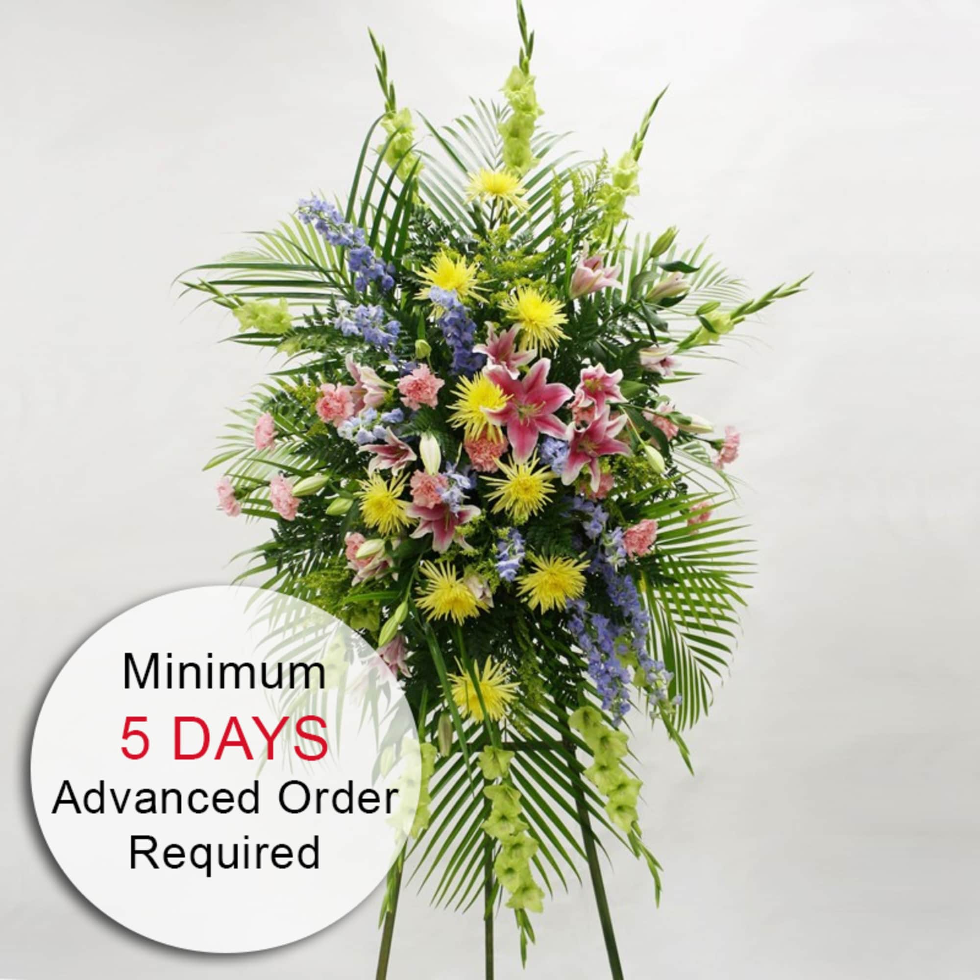Tall standing spray with pink lilies, yellow mums, blue delphinium, and pink carnations on an easel.
