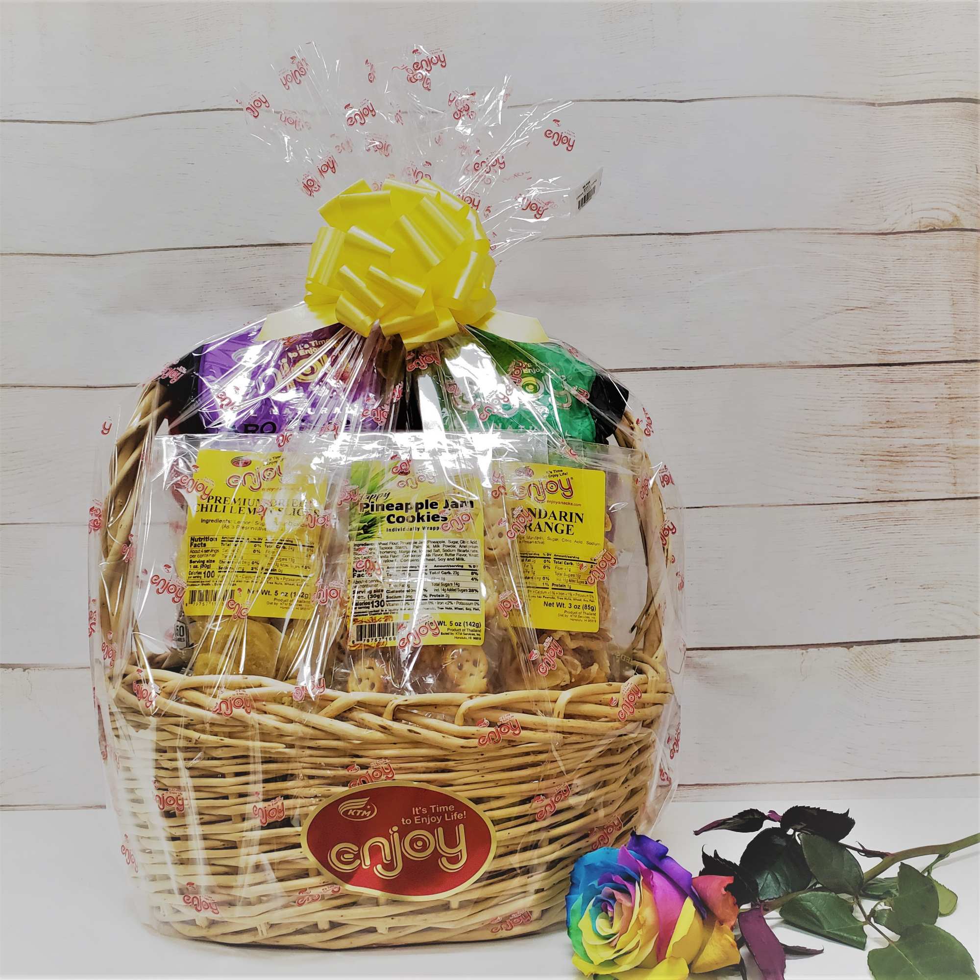 Wicker snack gift basket wrapped in cellophane with yellow bow and a single rainbow rose beside it