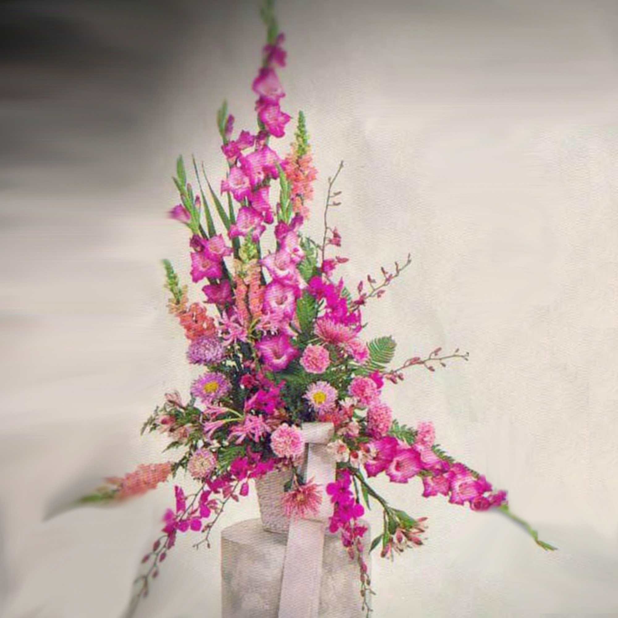 Tall pink floral arrangement with gladiolus-like stems and mums in a white container with a bow
