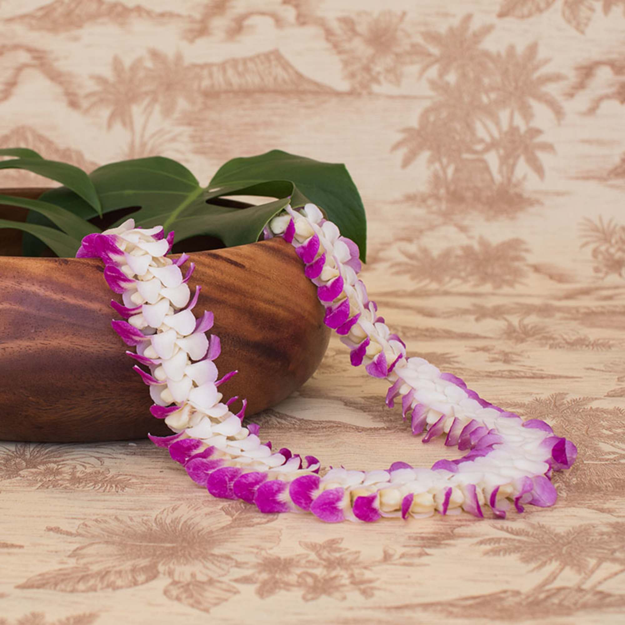 Purple and white orchid flower lei draped over a smooth wooden bowl with a tropical patterned background