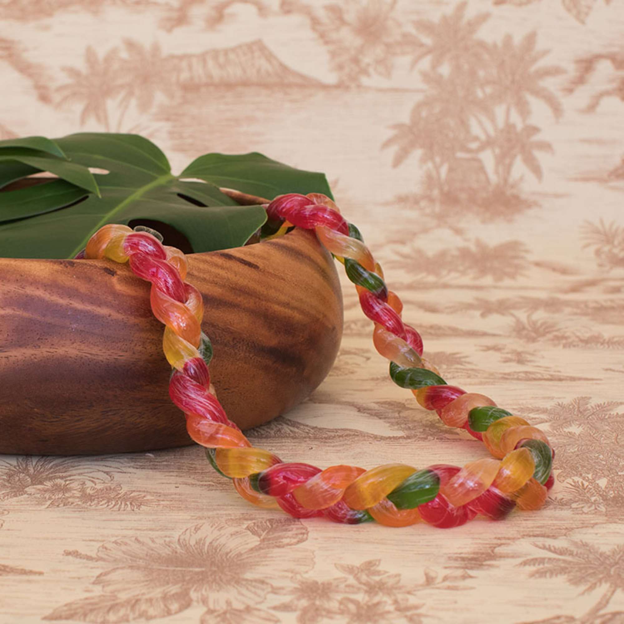 Twisted multicolor candy lei draped over a wooden bowl with a large tropical leaf