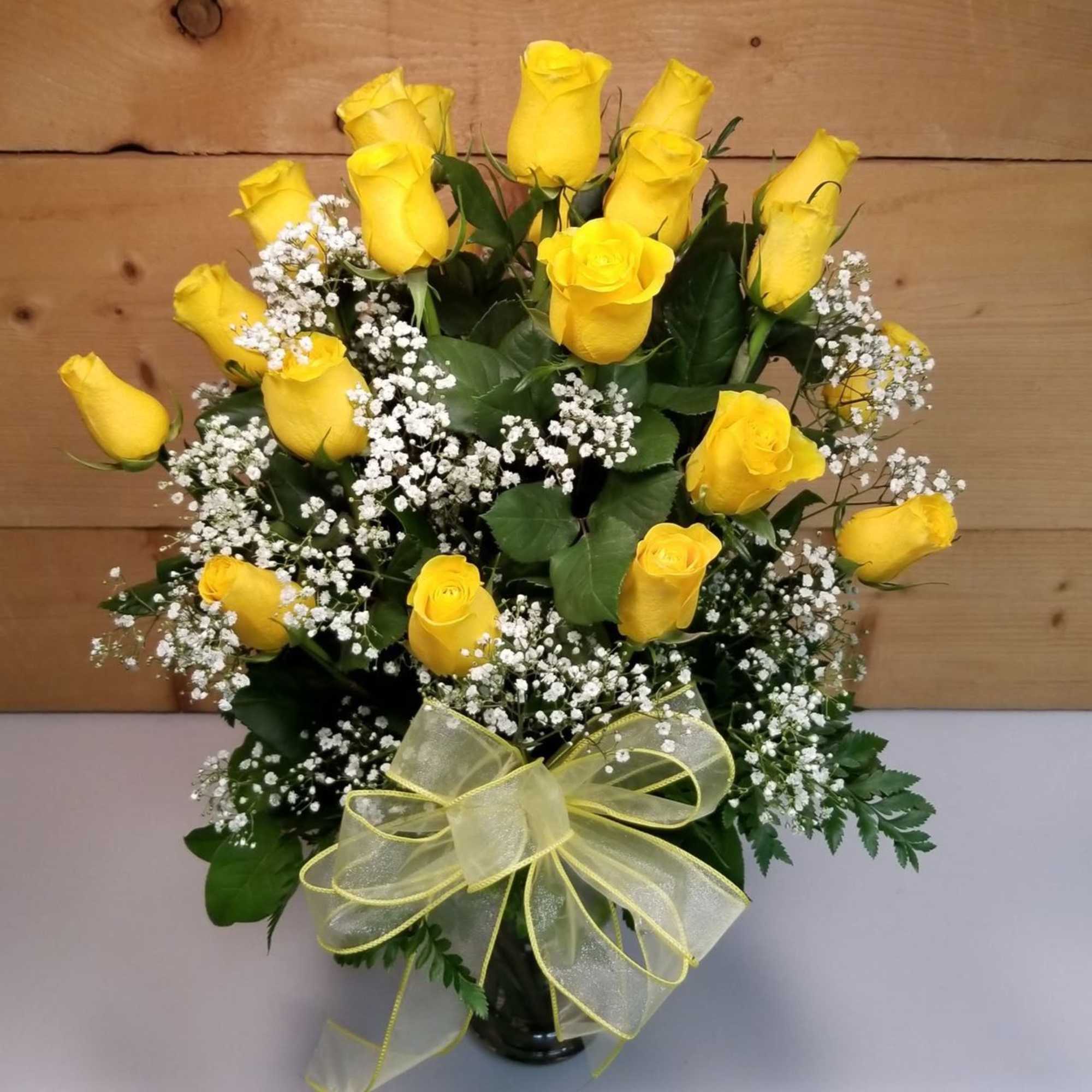 Tall arrangement of yellow roses and white filler flowers in a glass vase with a yellow bow