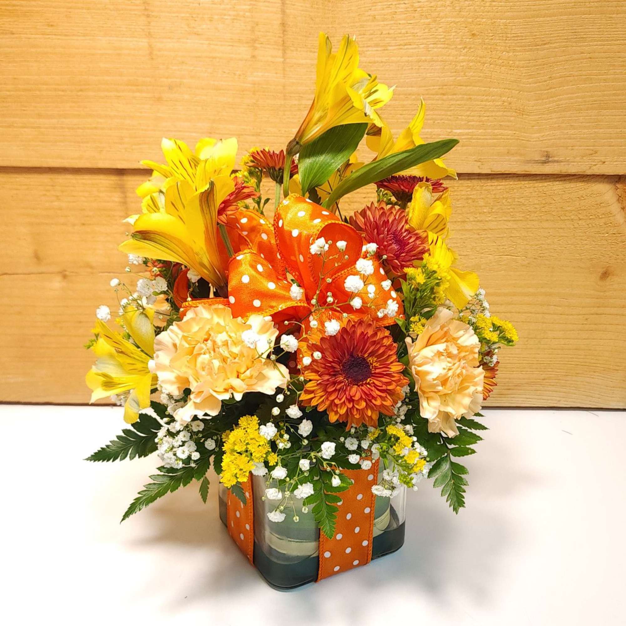 Mixed yellow and orange flower arrangement in a square glass vase with an orange ribbon
