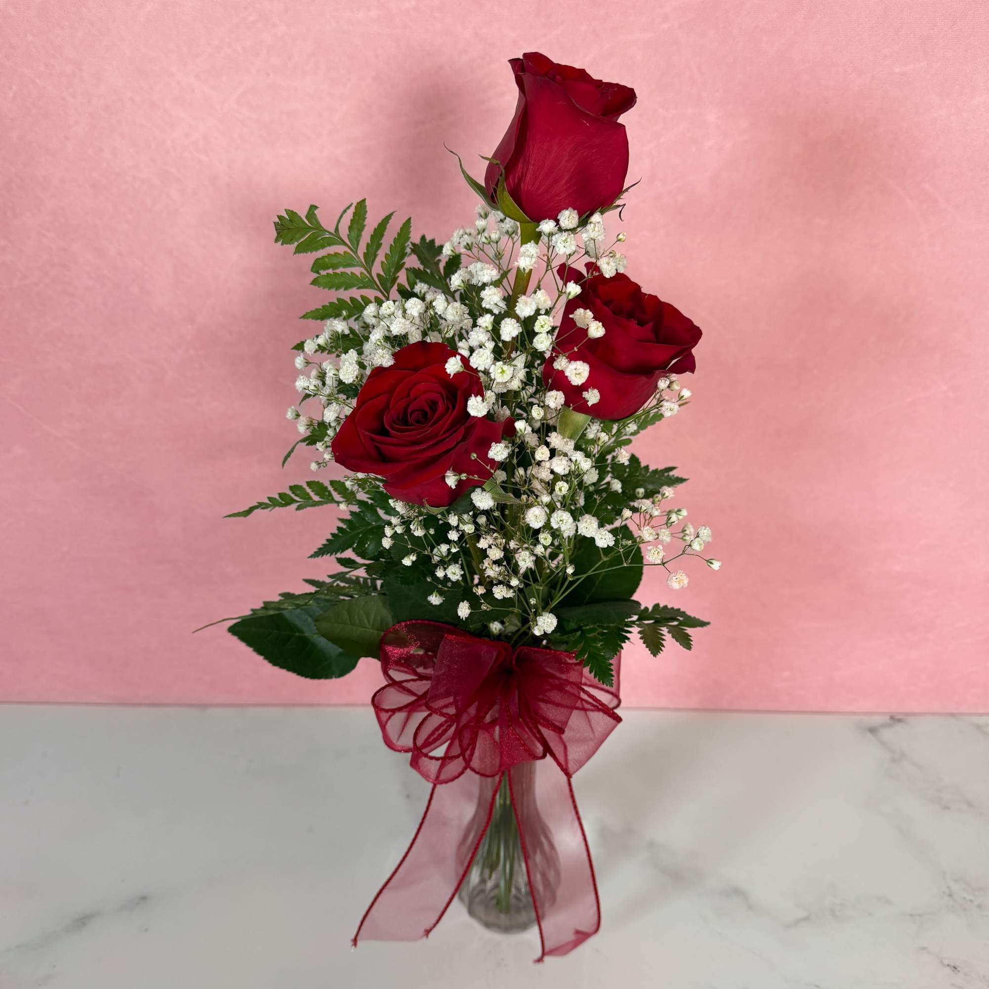Three red roses with baby's breath in a glass vase tied with a red ribbon.