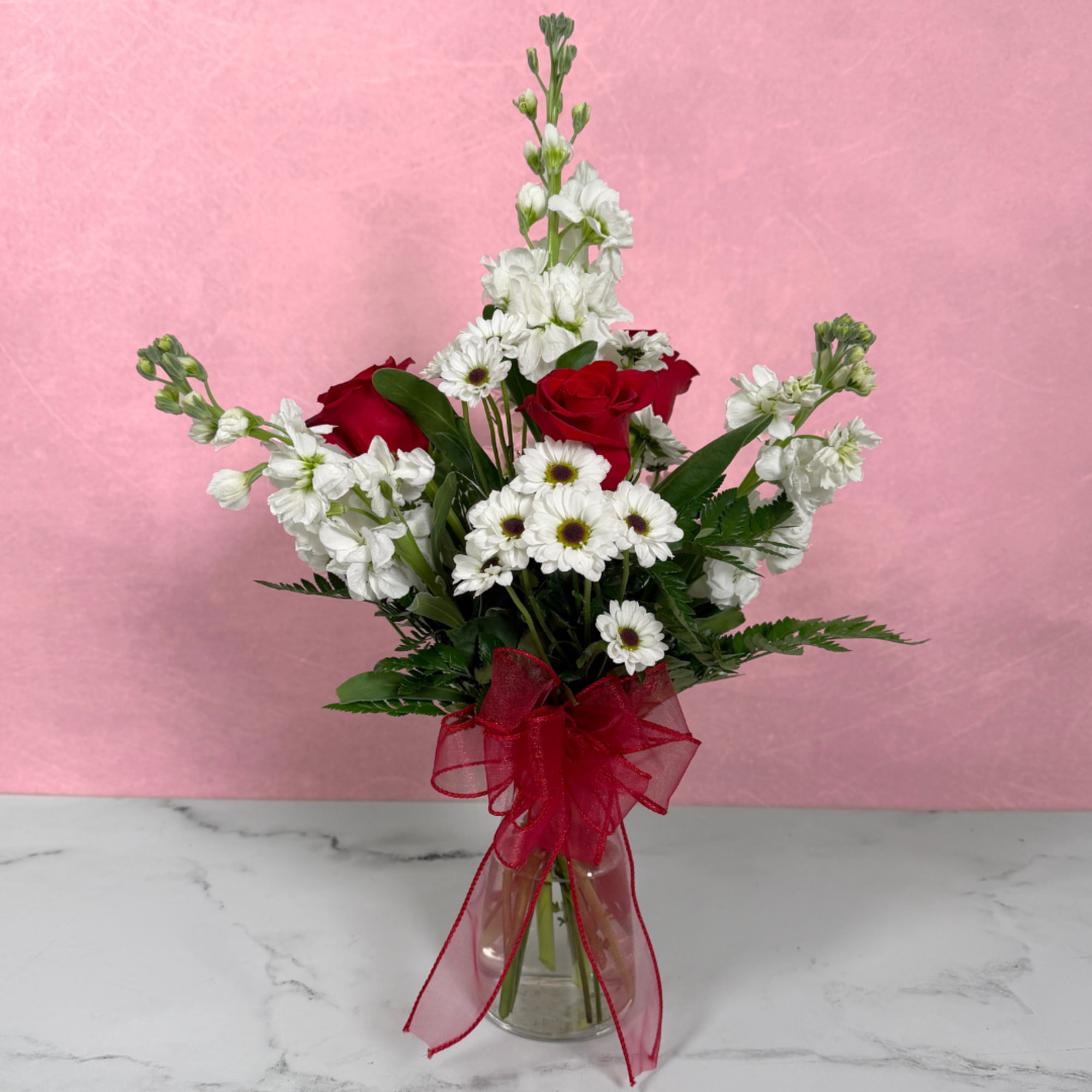Red roses and white daisies in a clear glass vase with a red ribbon