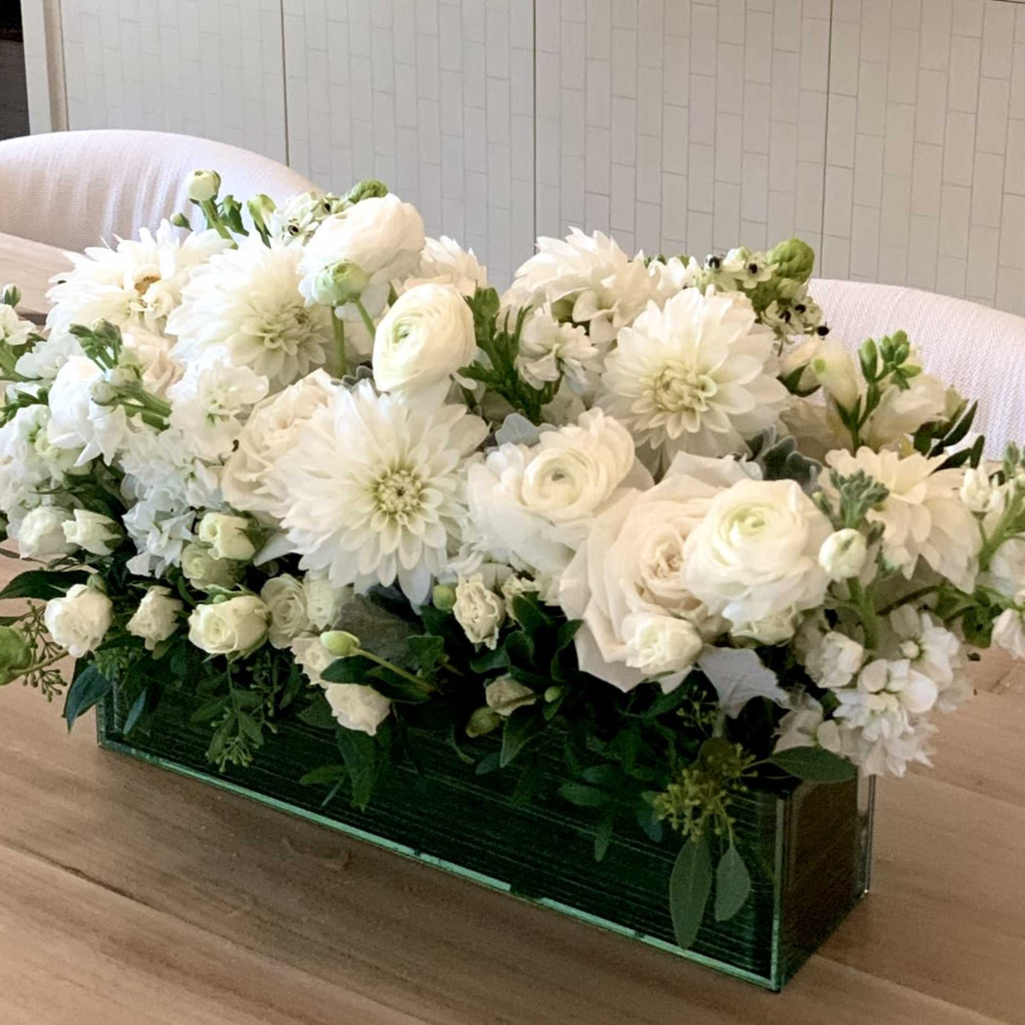 Low rectangular arrangement of white roses, dahlias, and other blooms in a green glass vase