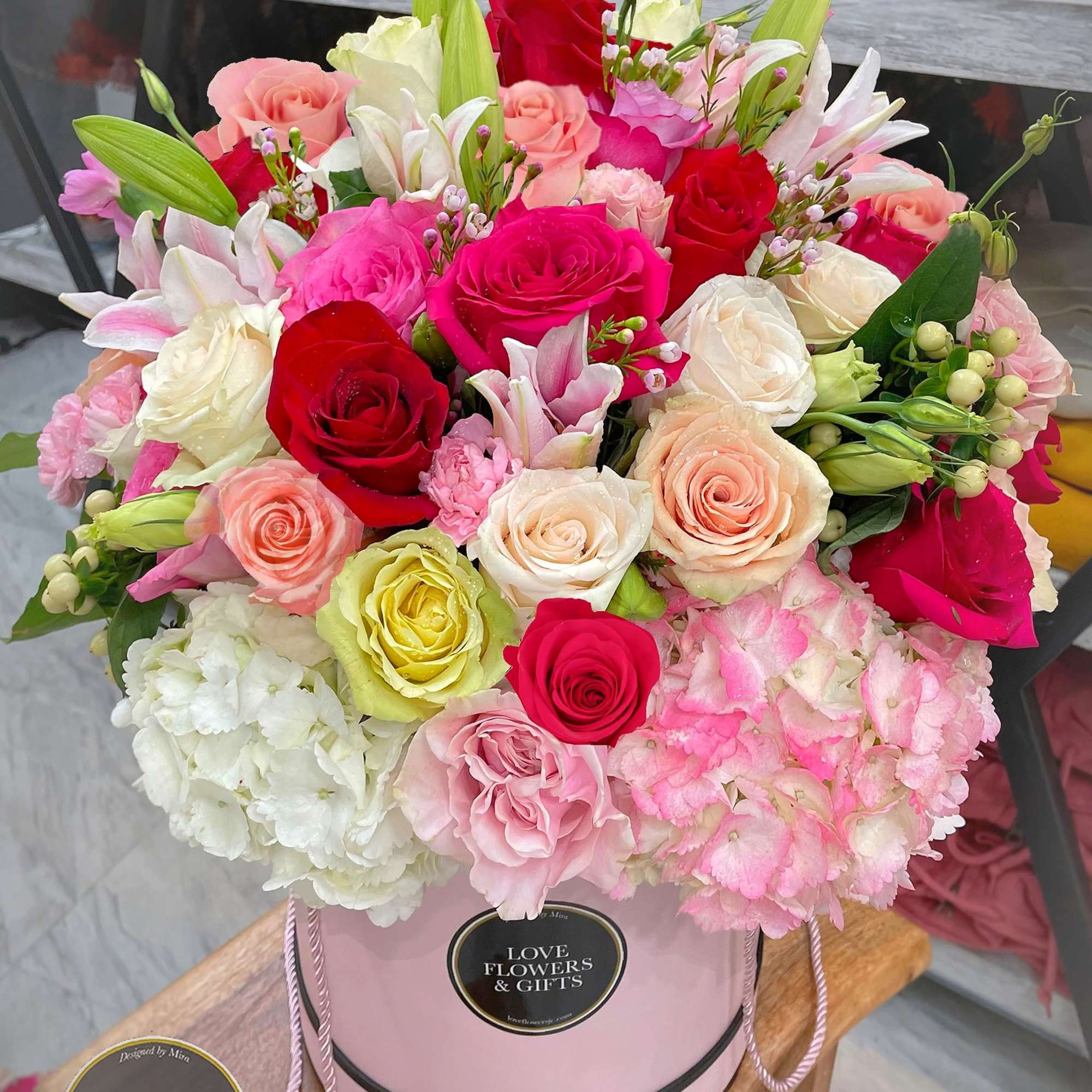 Round pink hatbox filled with mixed roses, lilies, and hydrangeas in red, pink, white, and peach tones