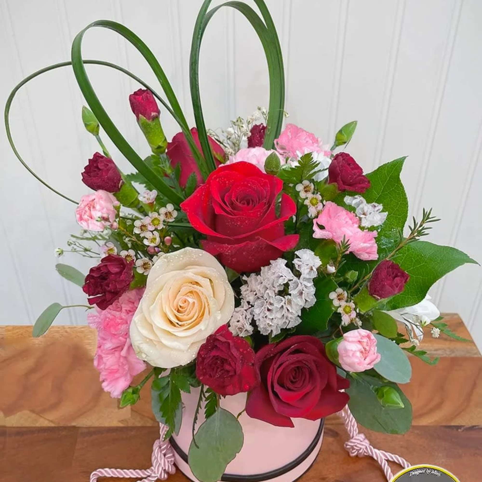 Pink hatbox arrangement with red and cream roses, pink carnations, and decorative grass loops on a wooden surface.