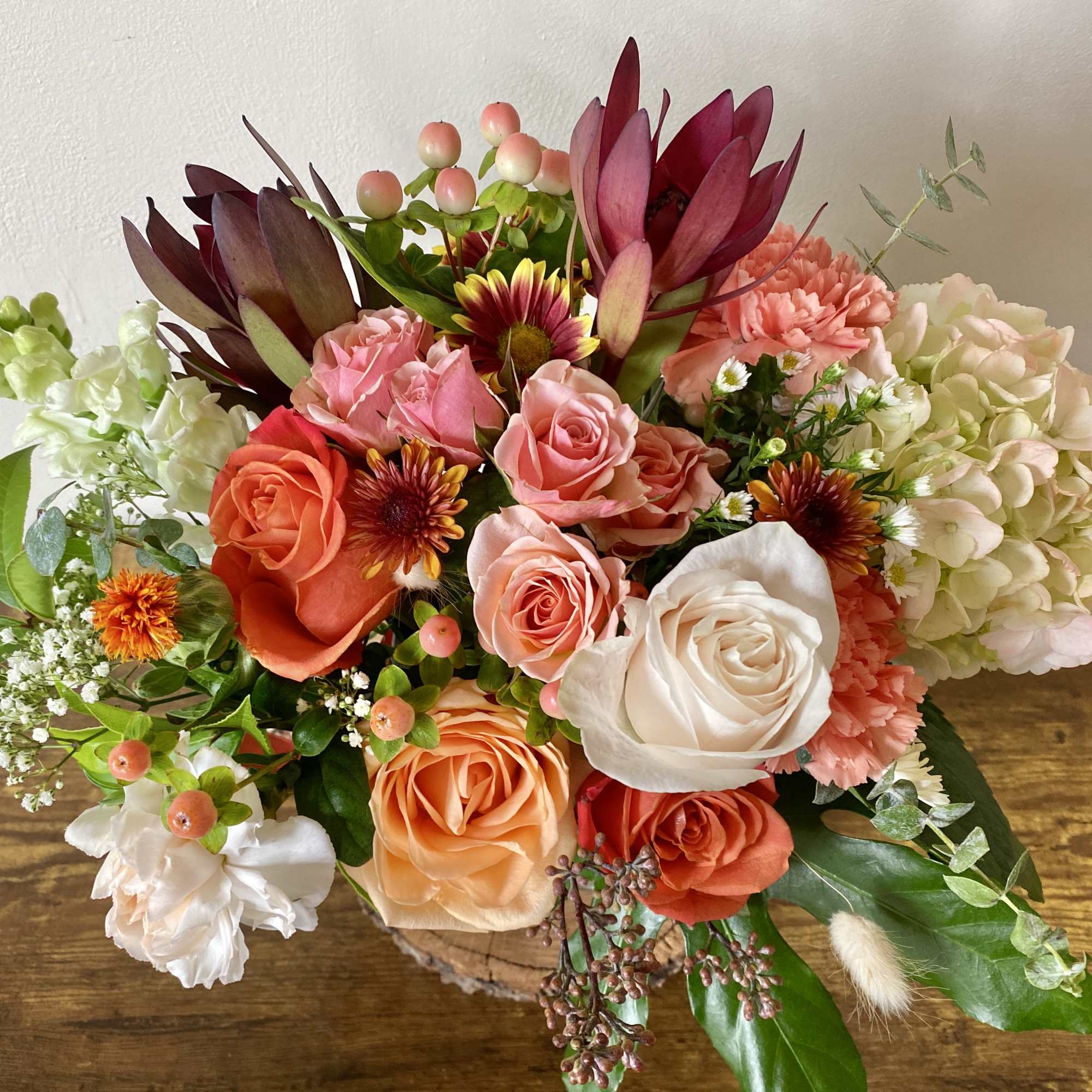 Mixed arrangement of peach and pink roses, carnations, hydrangea, and burgundy protea in a low container