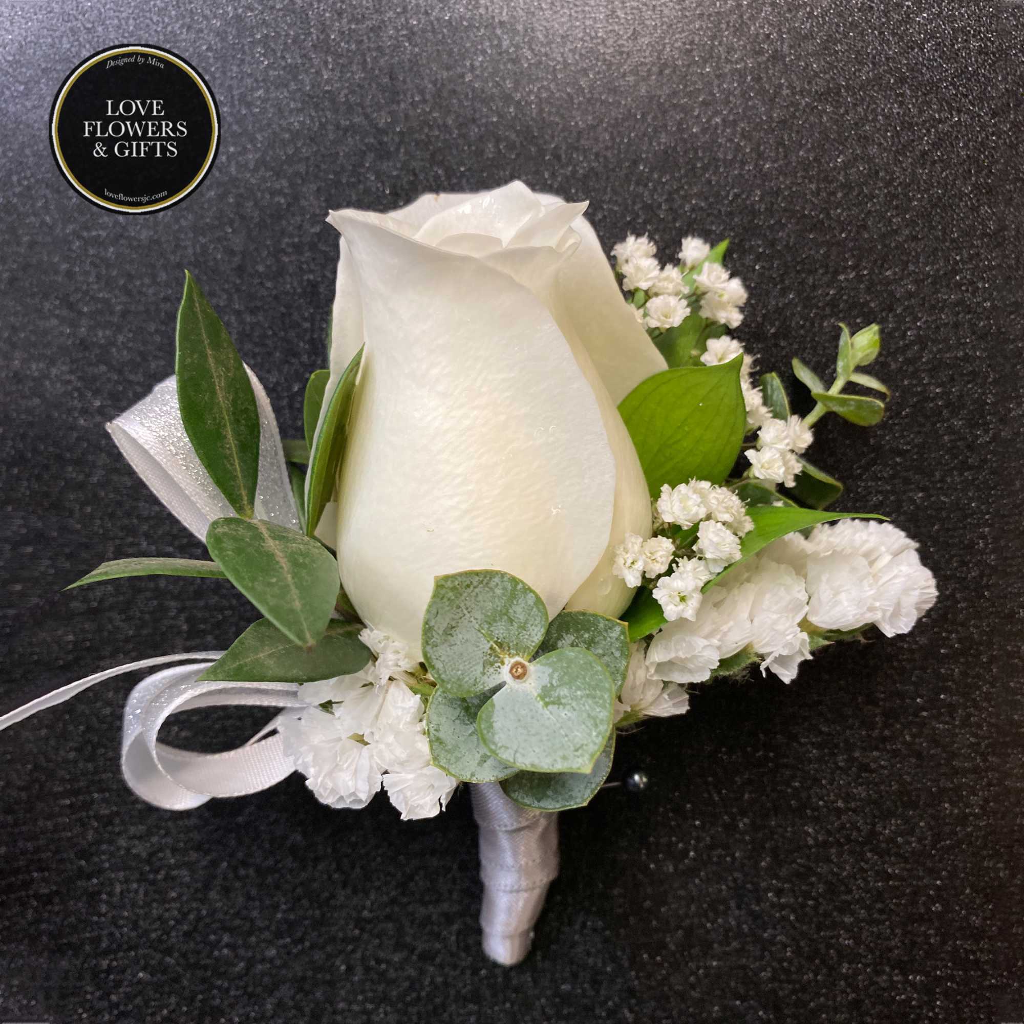 White rose boutonniere with small white filler flowers and silver ribbon on a dark background