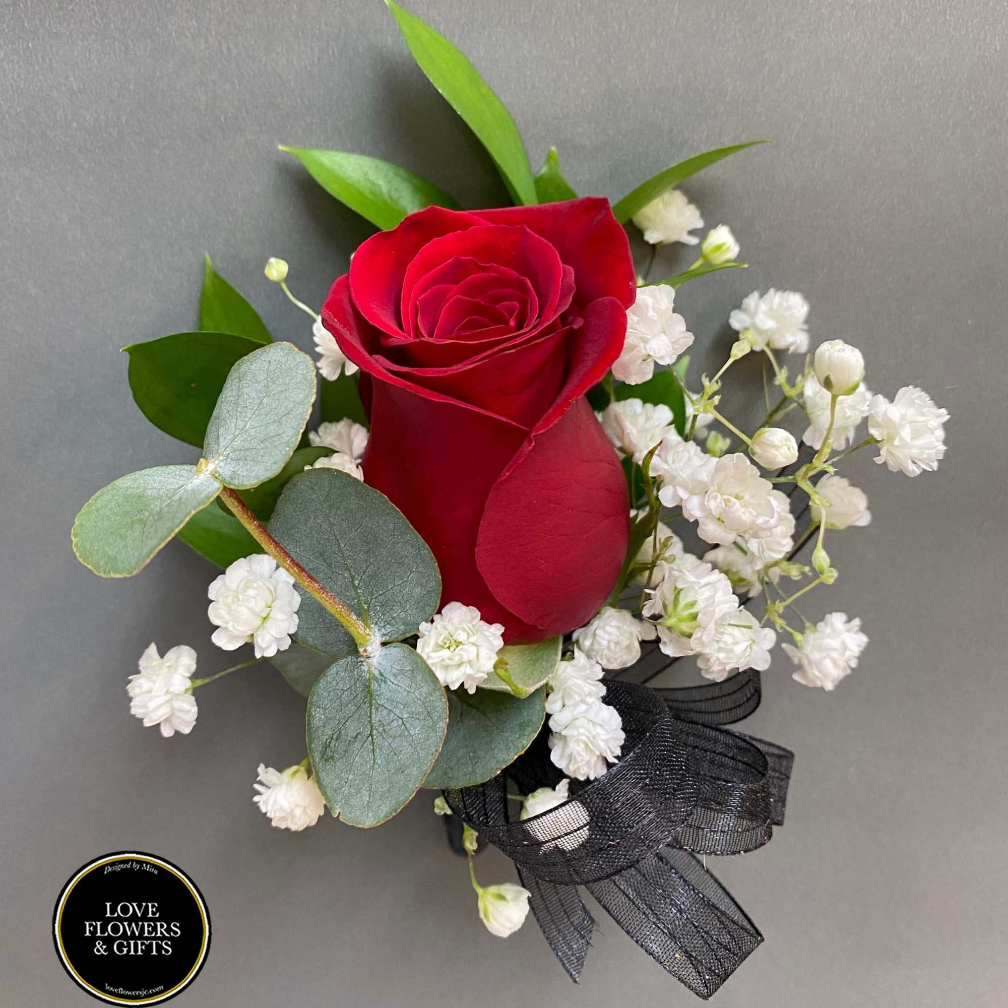 Red rose boutonniere with white filler flowers, eucalyptus leaves, and a black ribbon on a gray background