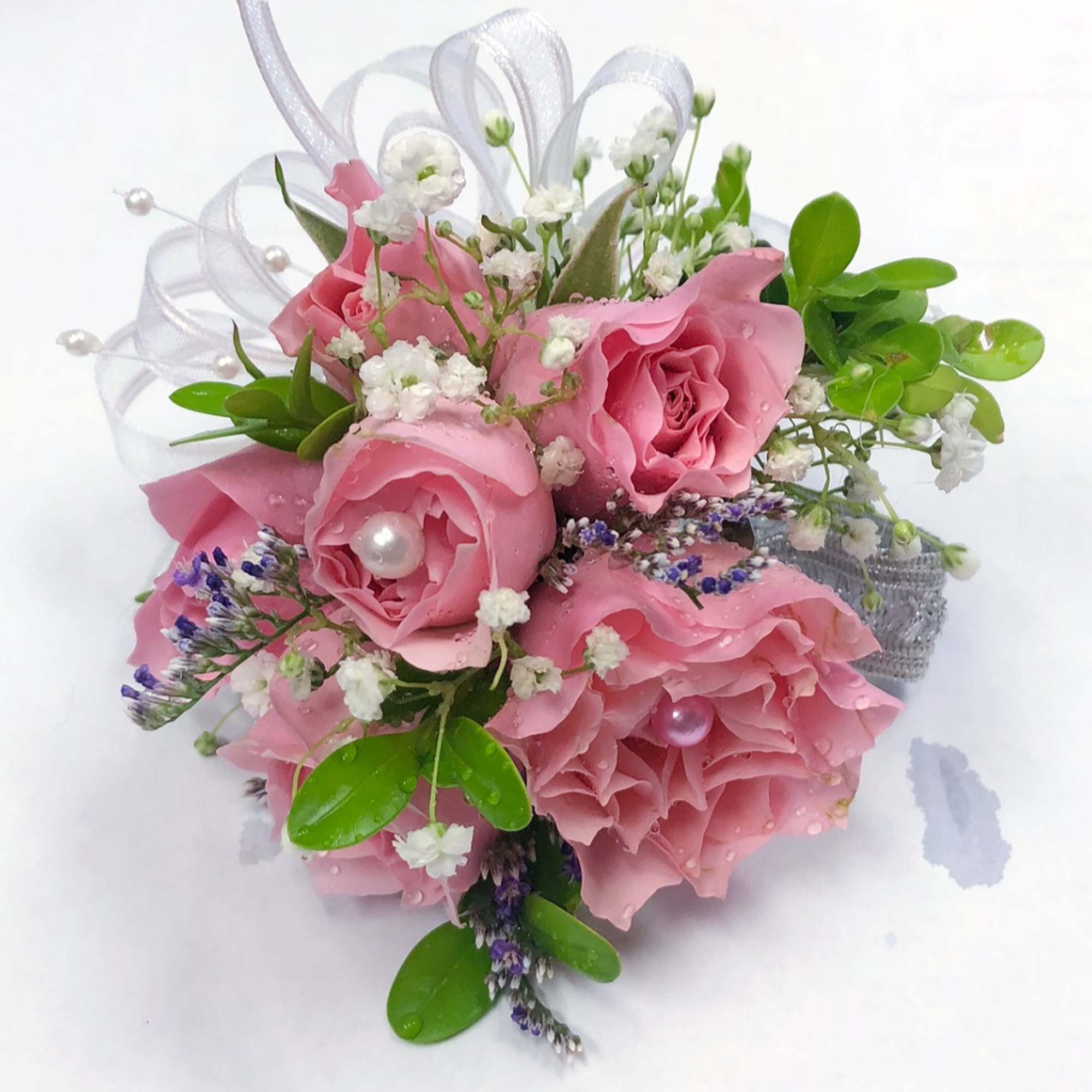 Pink rose wrist corsage with baby's breath, purple filler, greenery, ribbon loops, and pearl accents