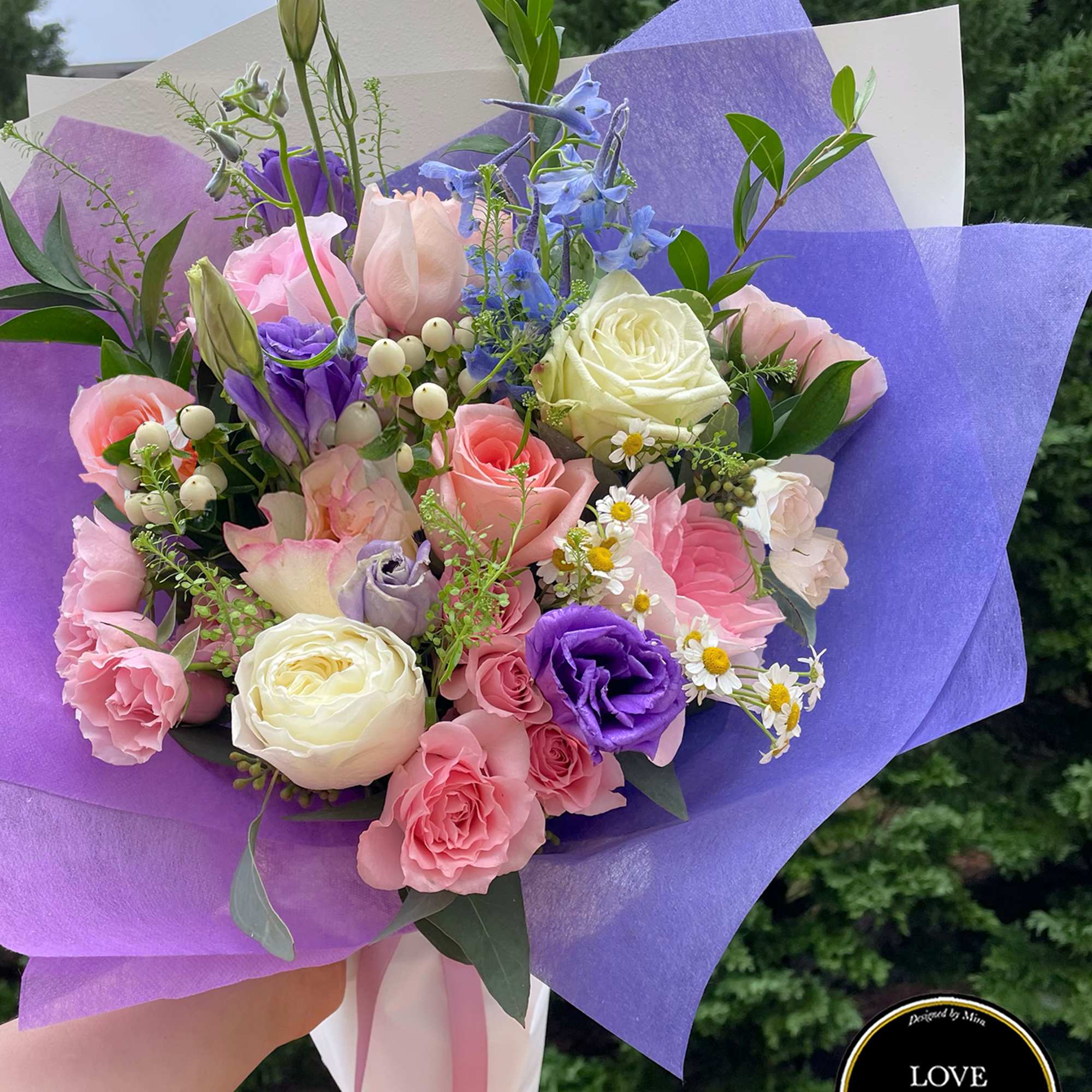 Hand-tied bouquet of pink roses, white blooms, and purple lisianthus wrapped in purple paper