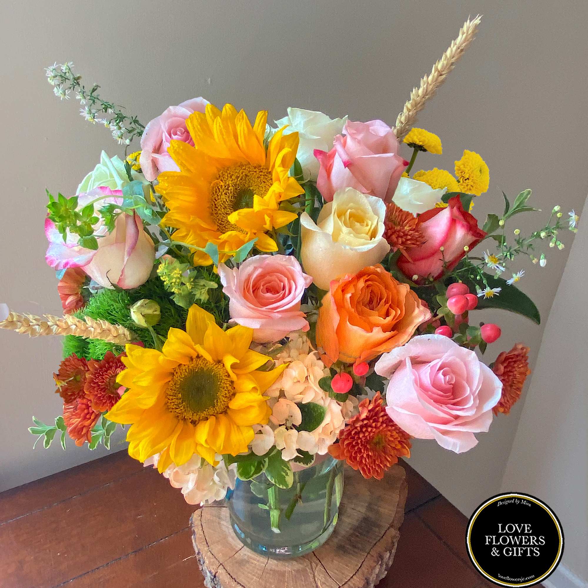 Mixed bouquet of sunflowers, roses, and mums in a clear glass vase on a wooden surface