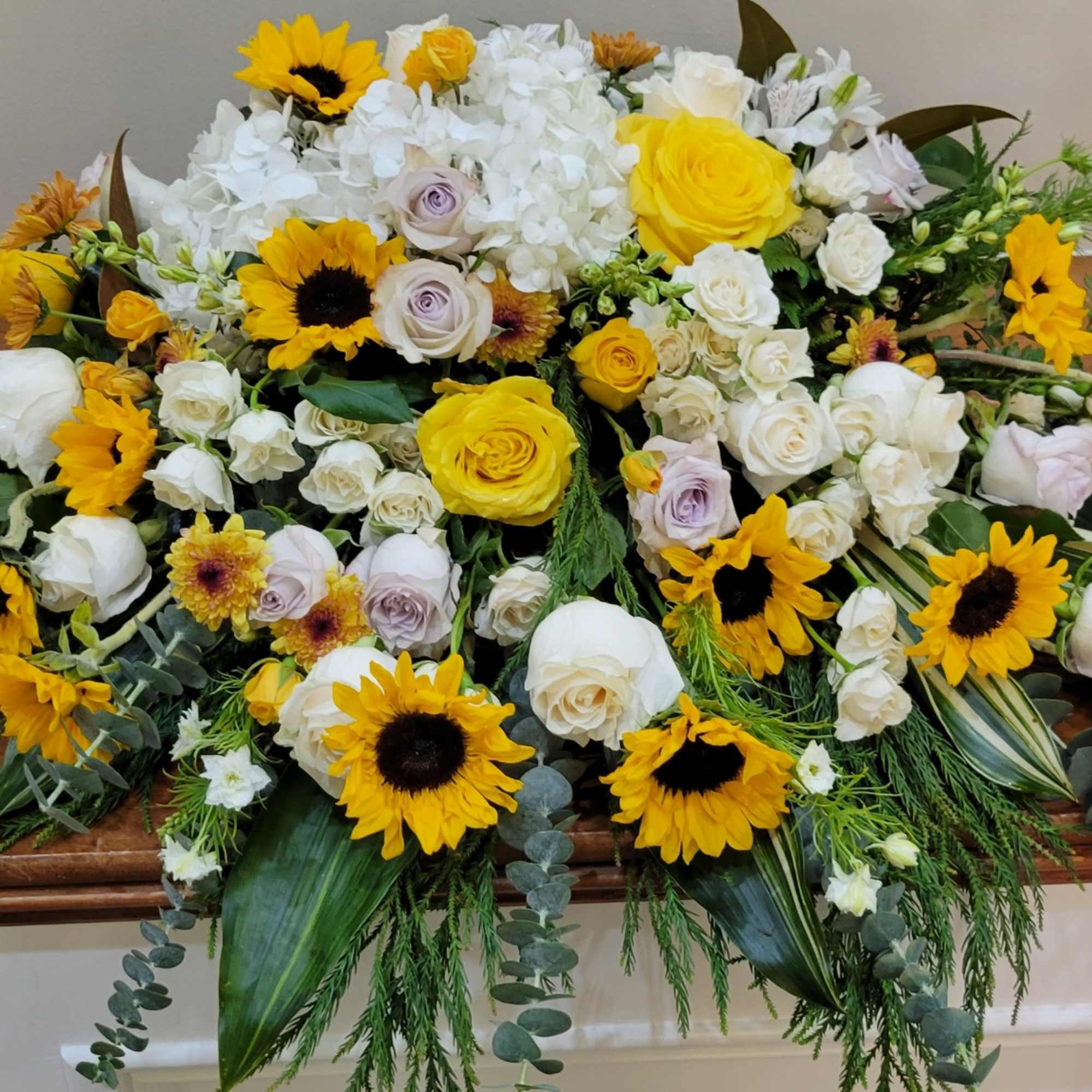 Casket spray of yellow sunflowers, white and yellow roses, and white hydrangeas on a wooden surface.