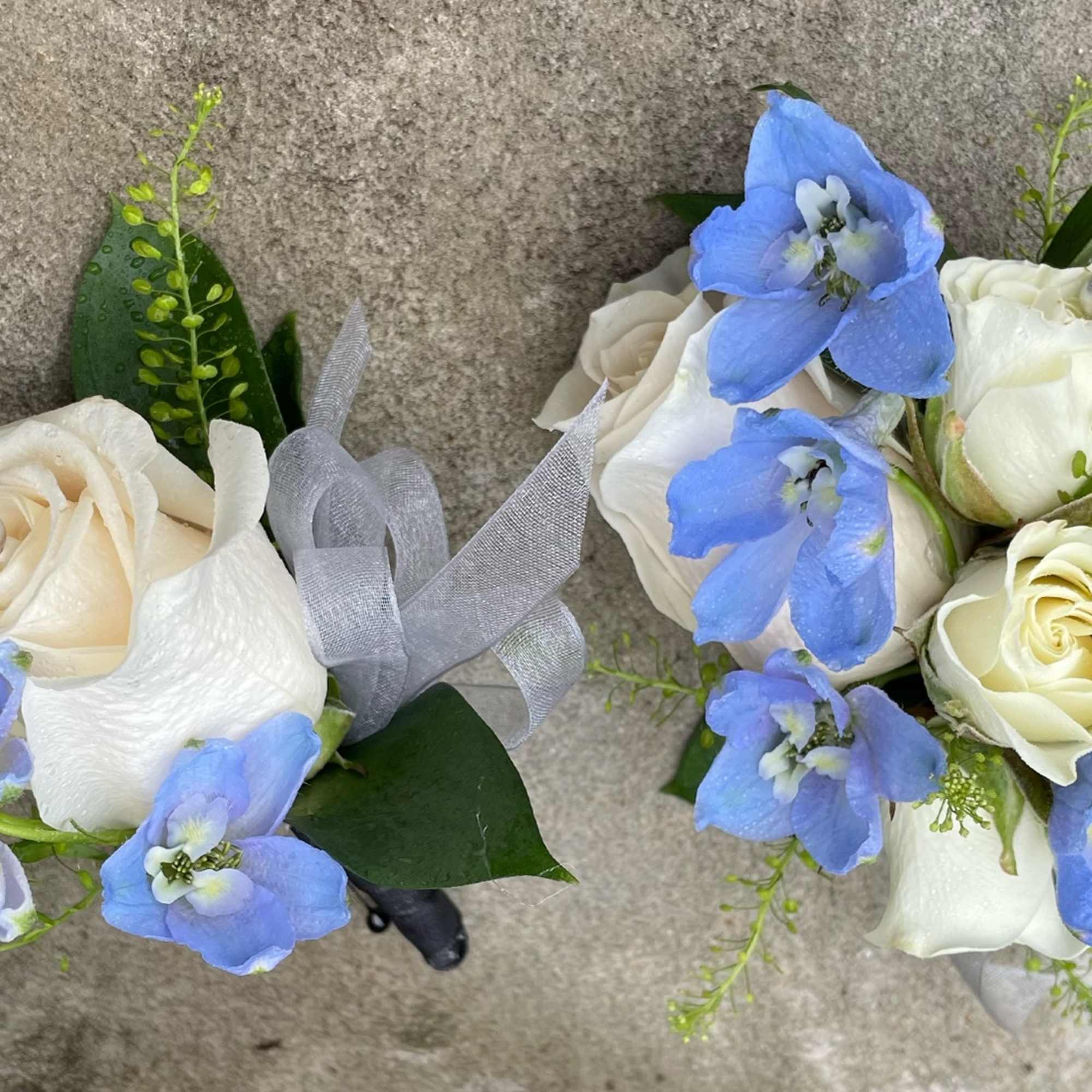 Pair of white rose and blue delphinium corsages with silver ribbon accents on a stone background