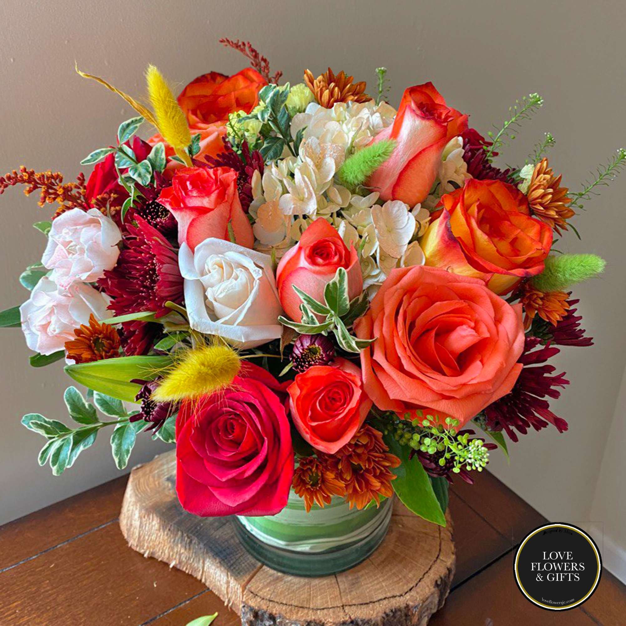 Vibrant arrangement of orange and red roses with white blooms in a glass vase on a wood slice.