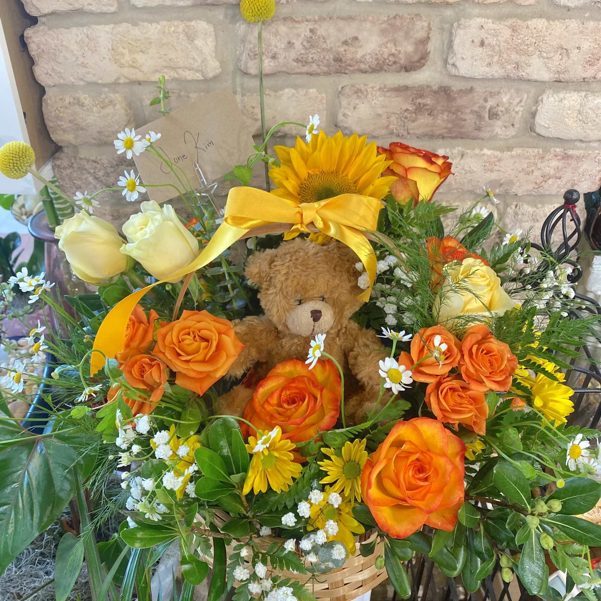 Basket of orange and yellow flowers with a small teddy bear and yellow bow in the center.