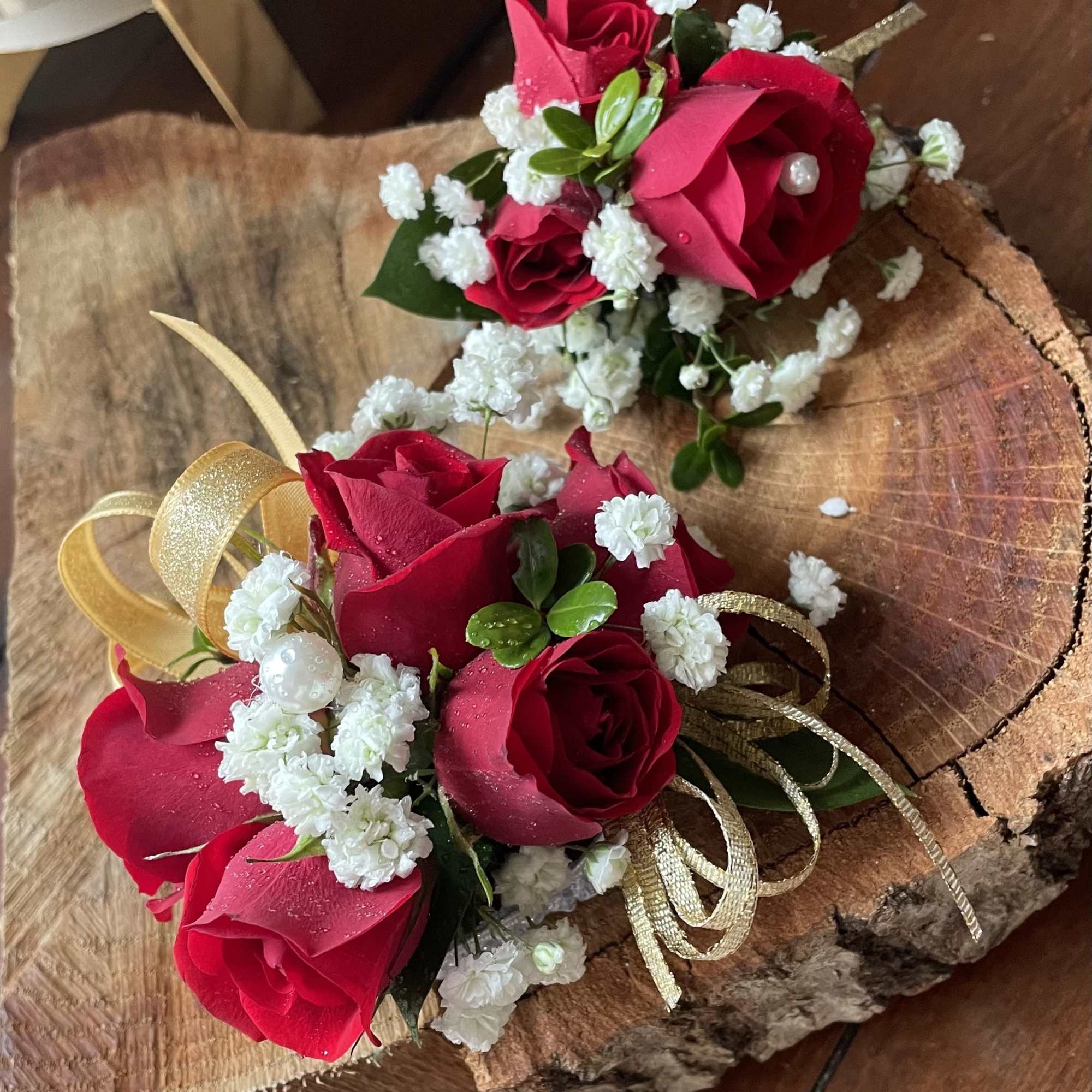 Two red rose corsages with white filler flowers and gold ribbon on a wood slab.
