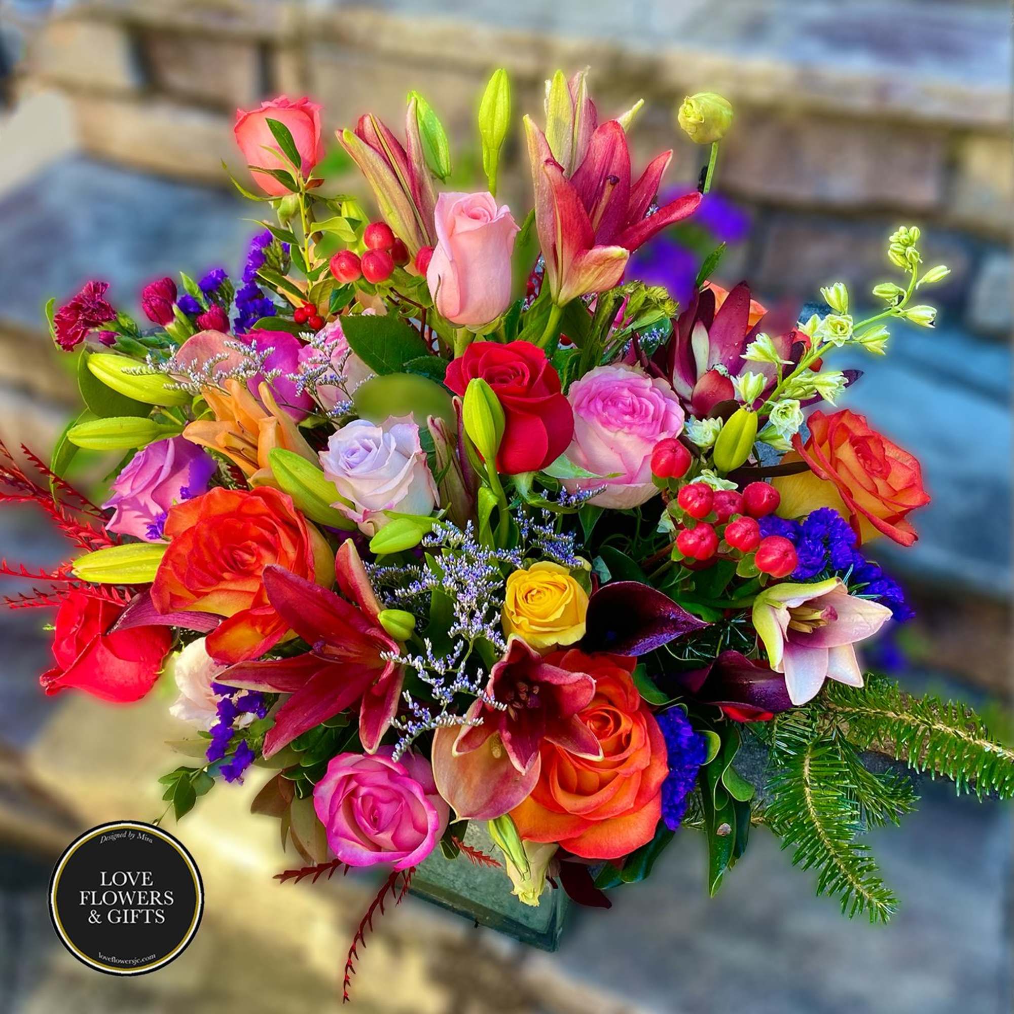 Vibrant mixed arrangement of roses and lilies in many colors arranged in a square glass vase.