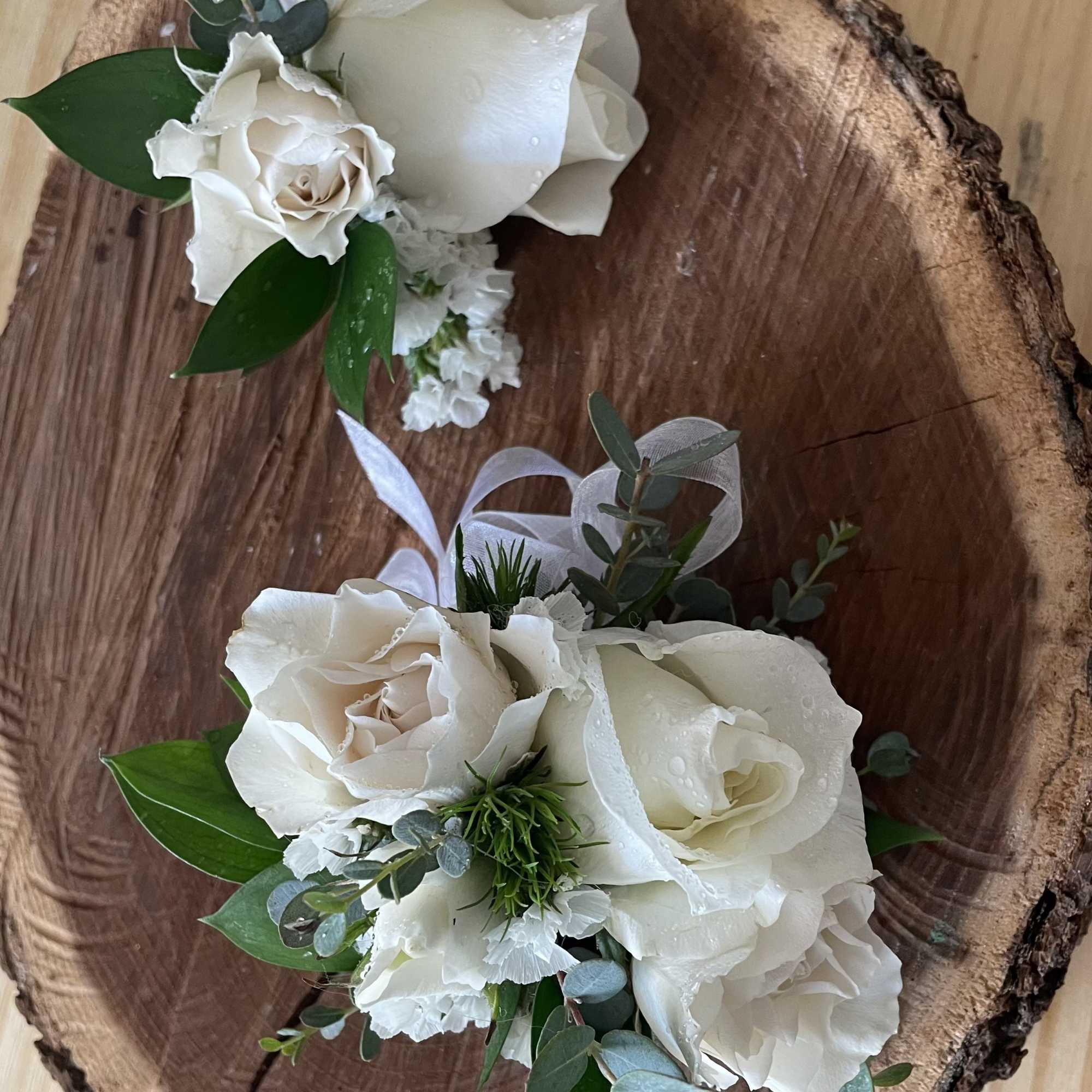 Two small white rose corsages with ribbon and greenery on a wood slice.