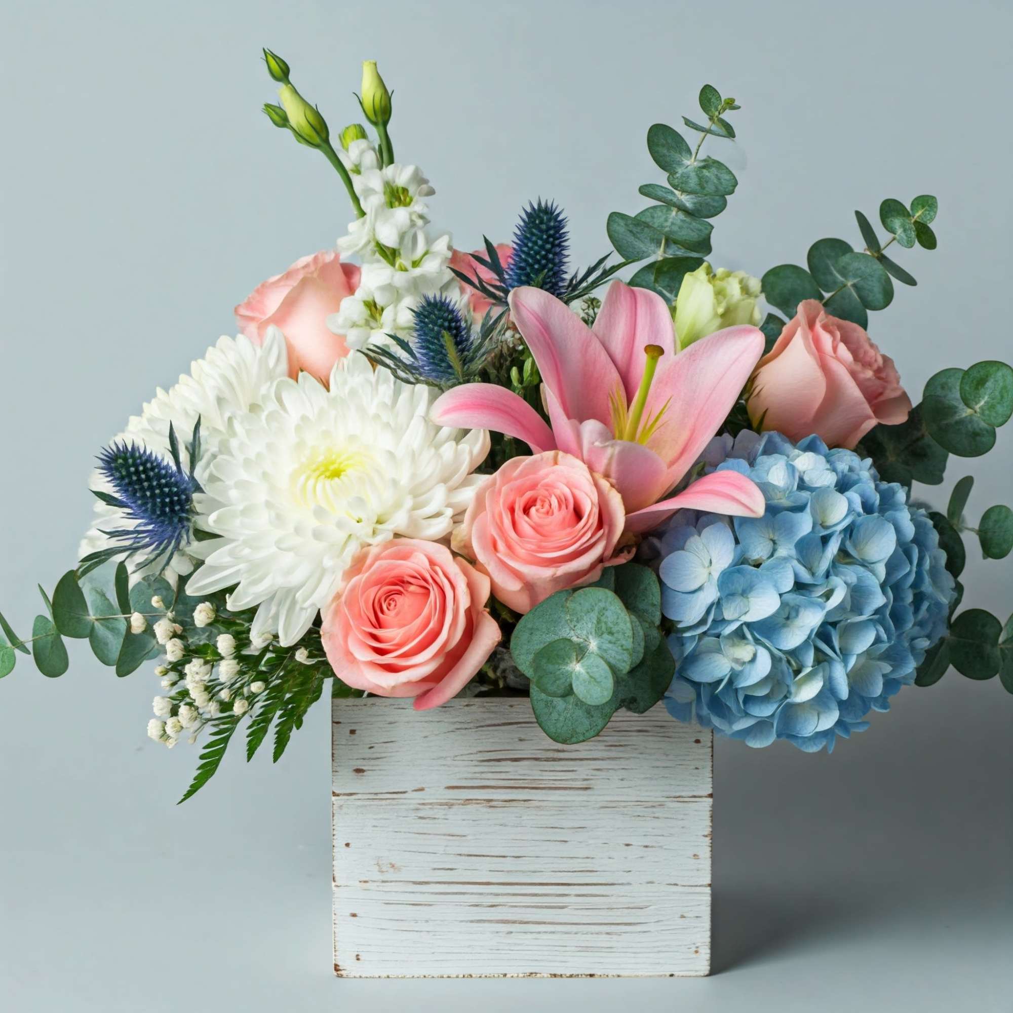 Pink roses, white blooms, blue hydrangea, and lilies in a white wooden box