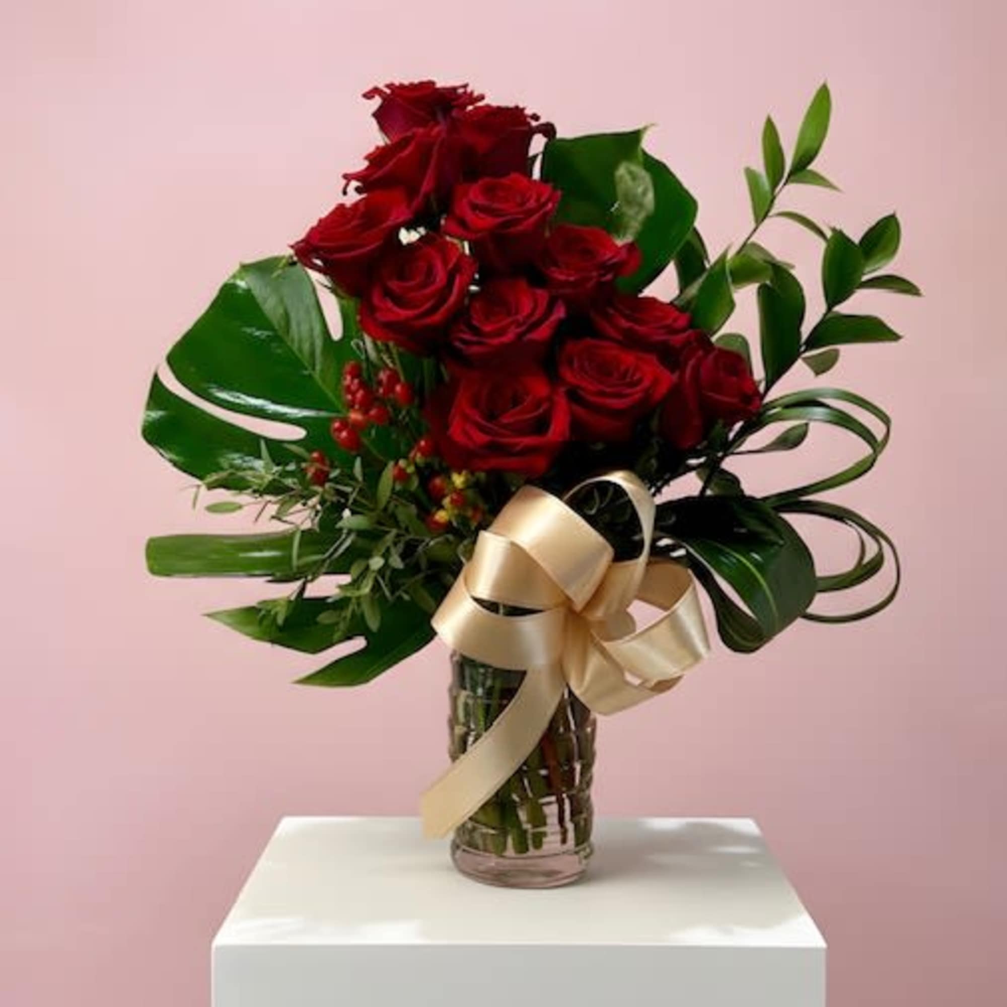 Tall arrangement of red roses with large green leaves in a glass vase with a gold bow
