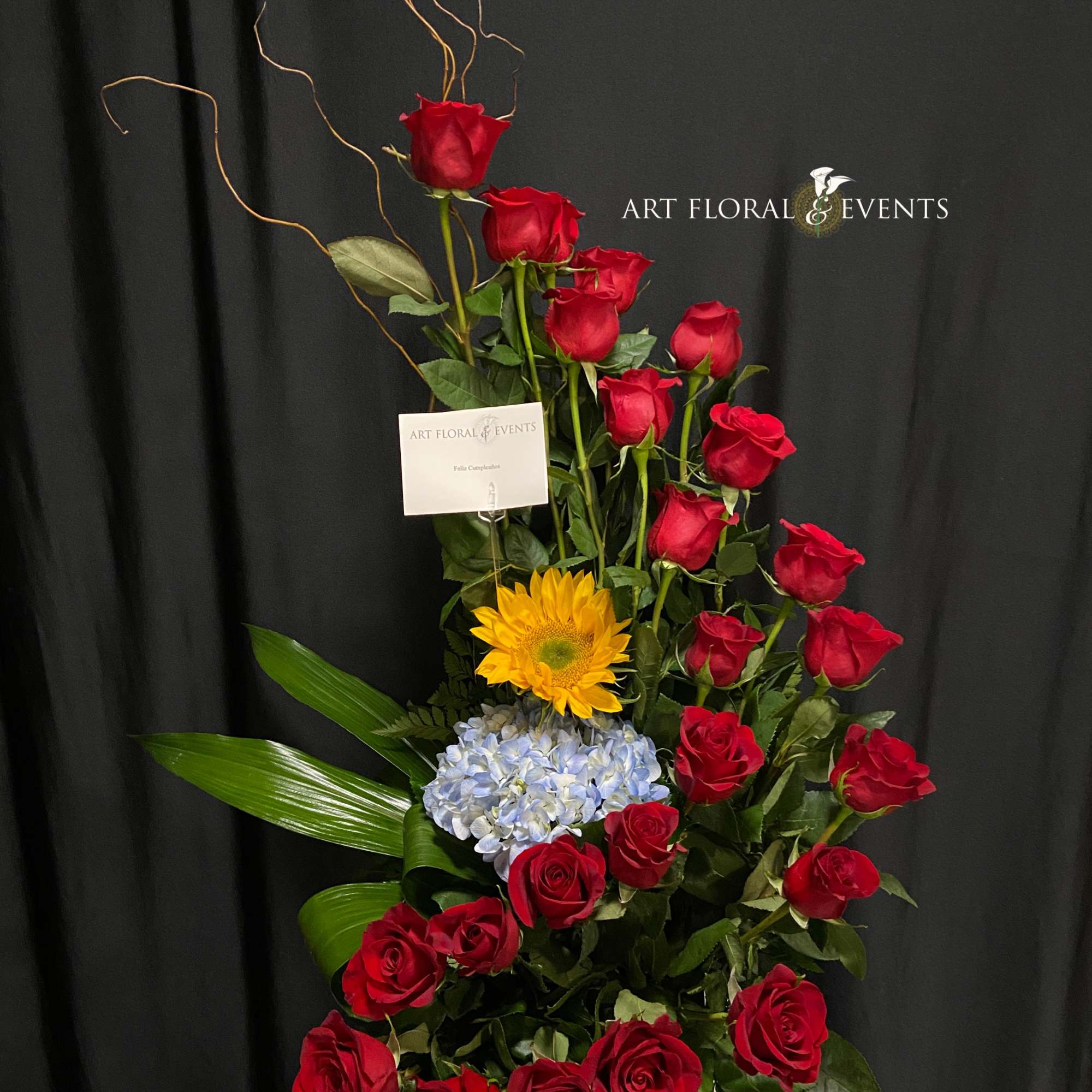 Tall arrangement of red roses with a yellow sunflower and blue hydrangea in a white vase