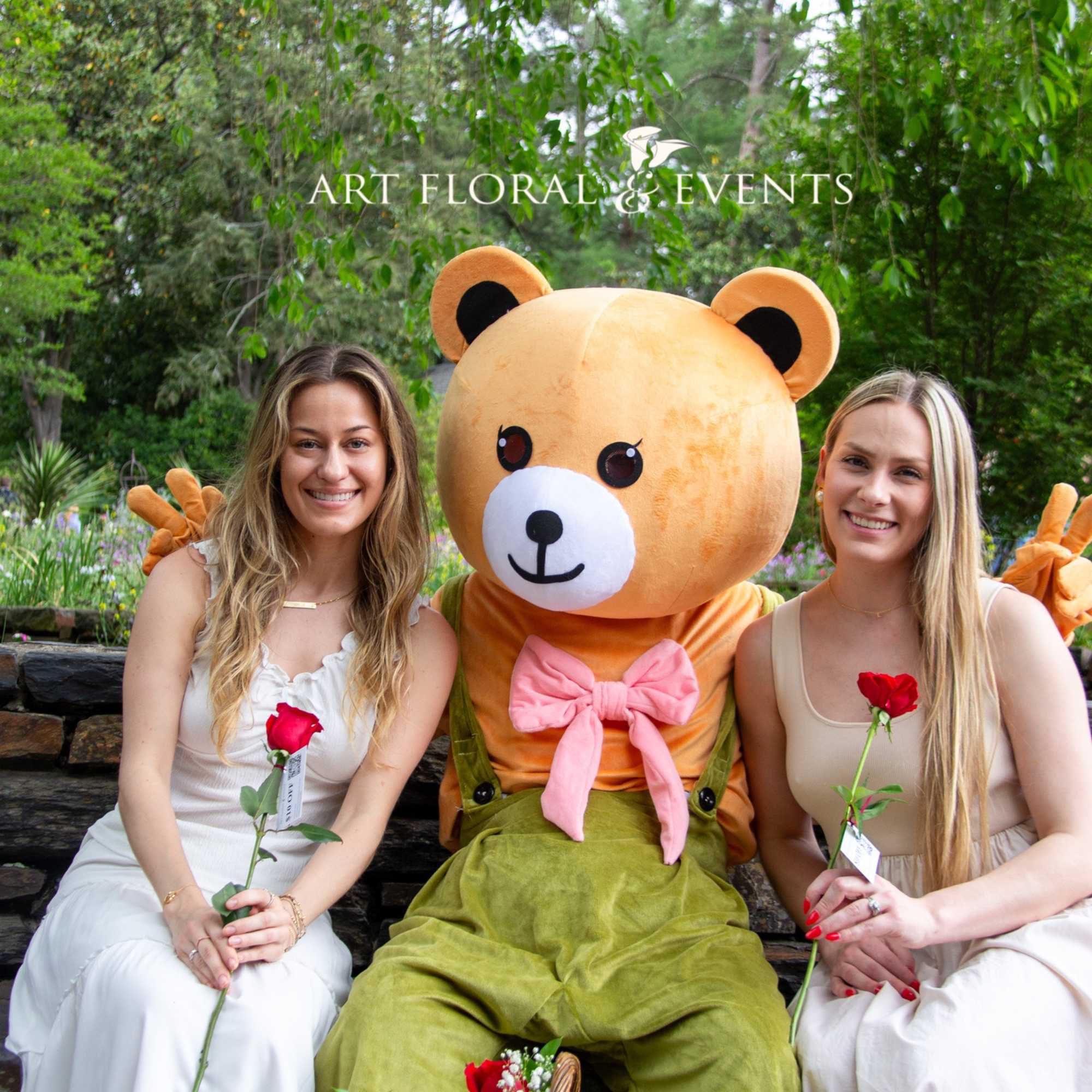 Two women sit beside a large teddy bear mascot holding red roses.