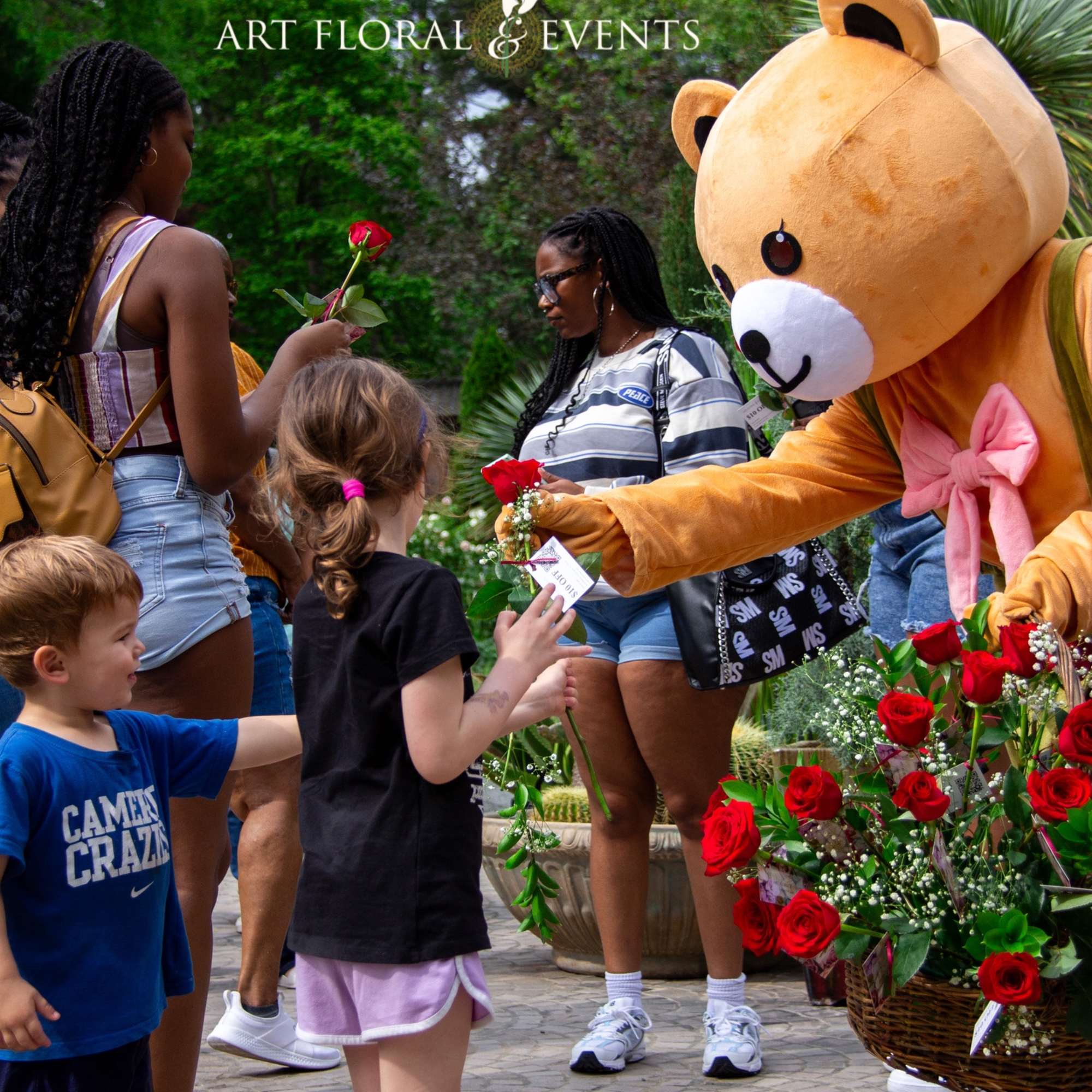 Children receive red roses from a teddy bear mascot beside floral baskets.