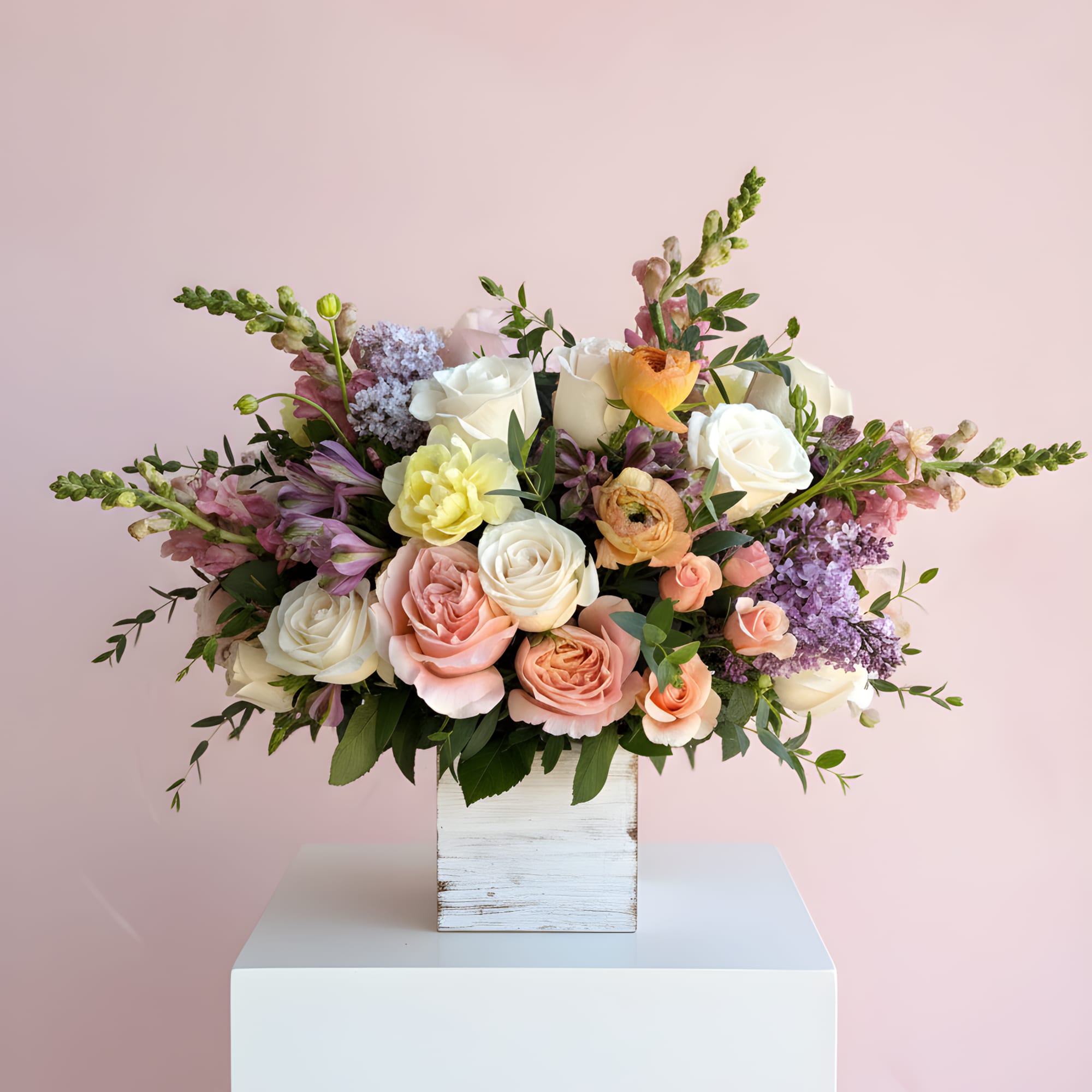 Low pastel arrangement of roses, ranunculus, lilac, and mixed blooms in a whitewashed wood box against a pink background
