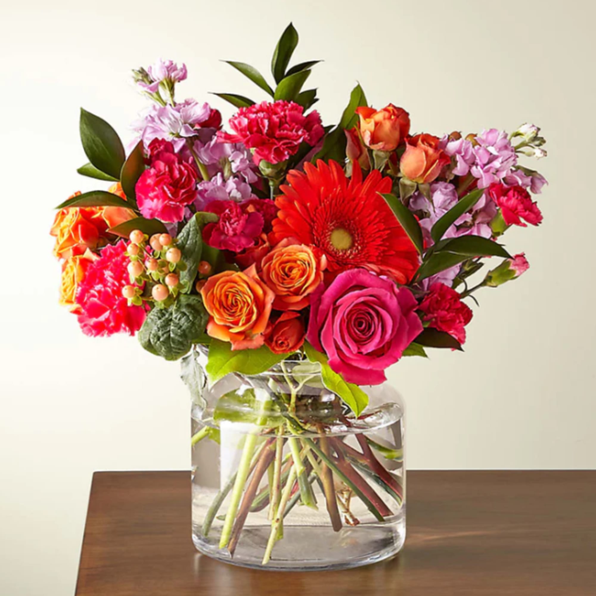 Bright mixed bouquet of roses, carnations, and gerbera daisies in a clear glass vase.