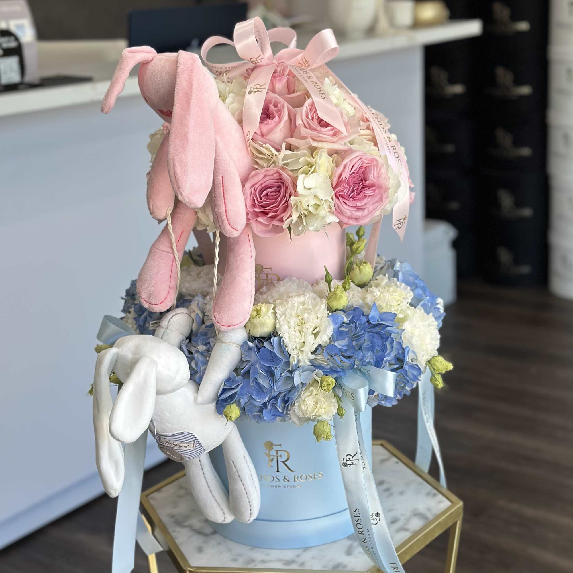Pink roses and blue hydrangeas in a pastel hatbox with plush bunnies