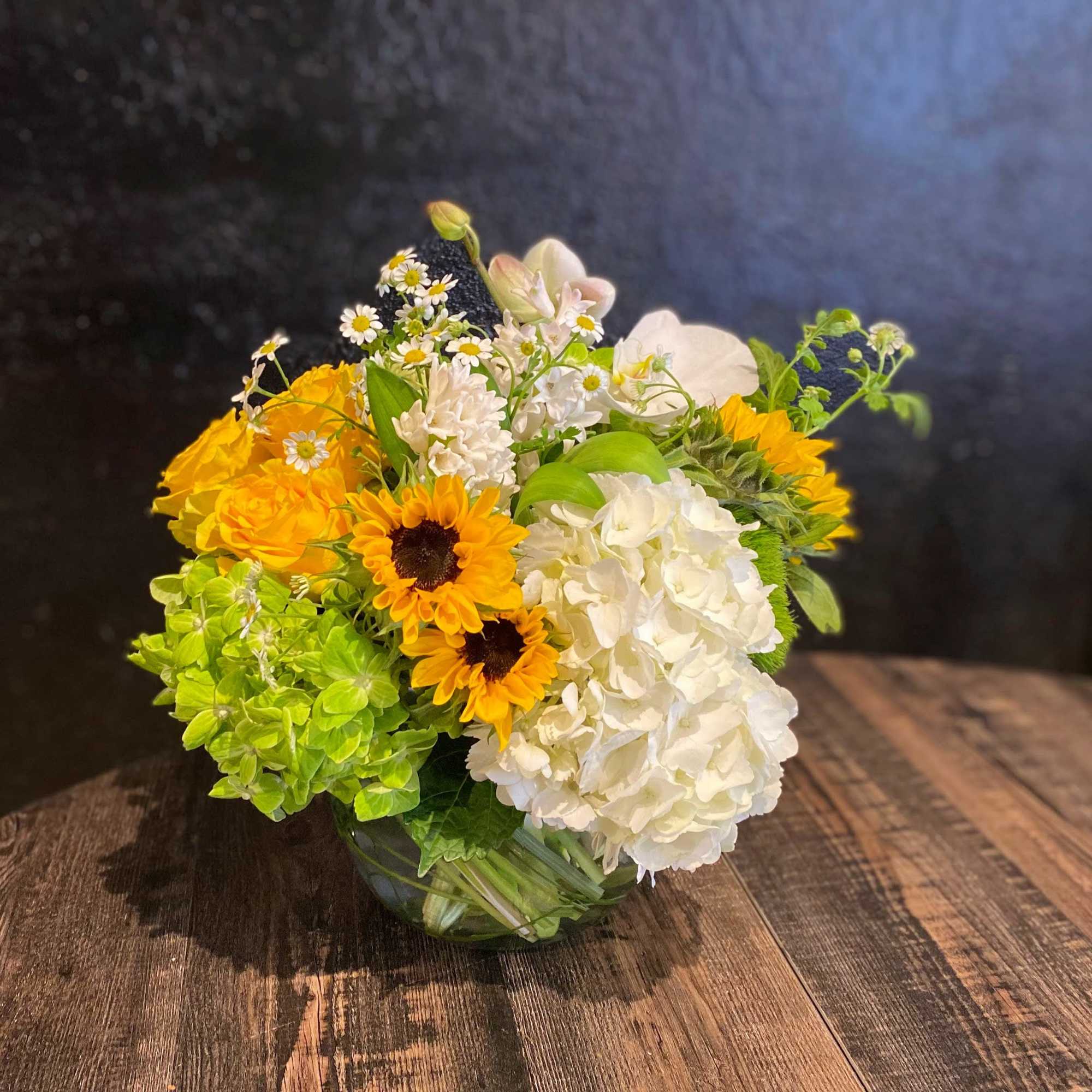 Low mixed arrangement of yellow sunflowers, white hydrangeas, and bright green blooms in a clear glass bowl.