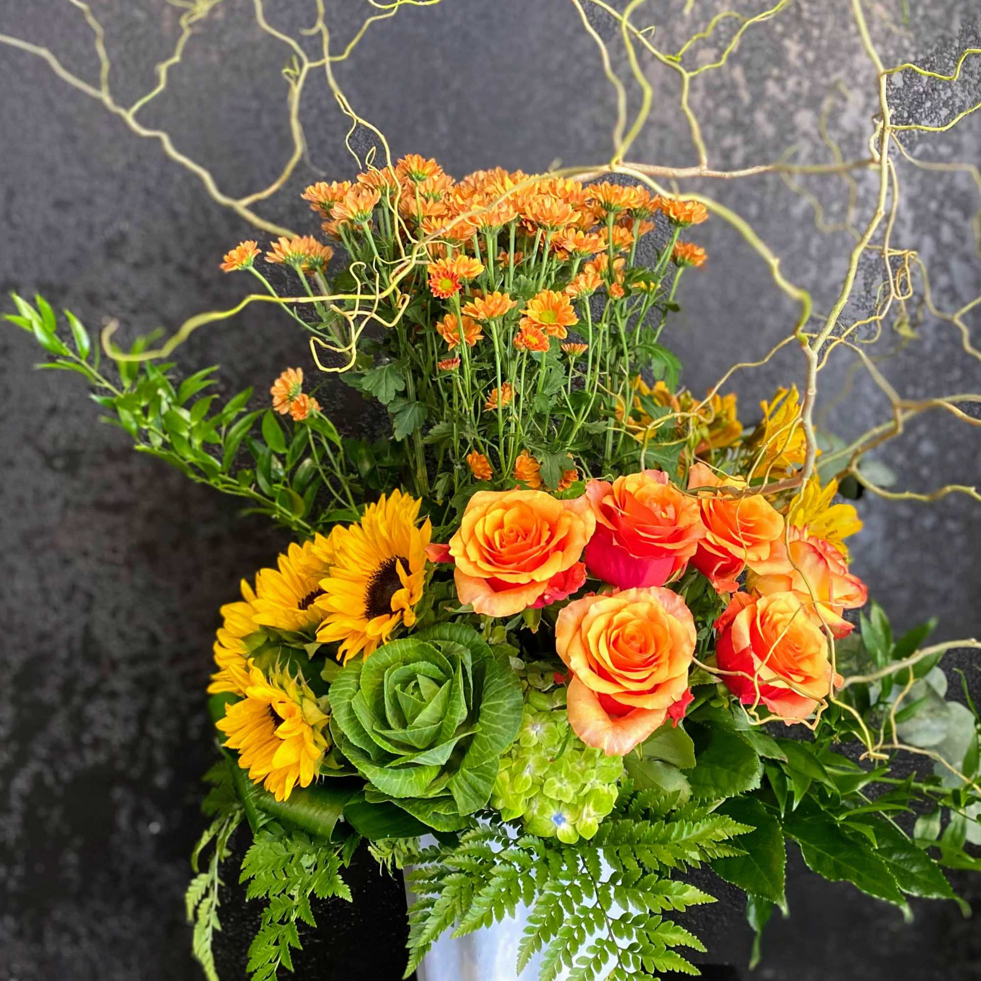 Tall orange and yellow arrangement with roses, sunflowers and mums in a white vase with curly branches
