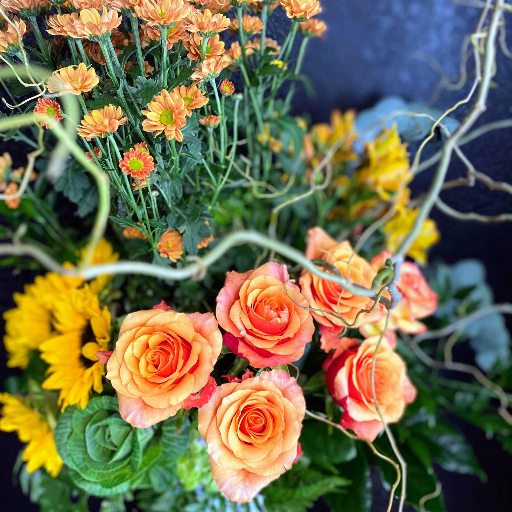 Tall arrangement of orange roses, yellow sunflowers, and orange mums with curly branches in a dark container