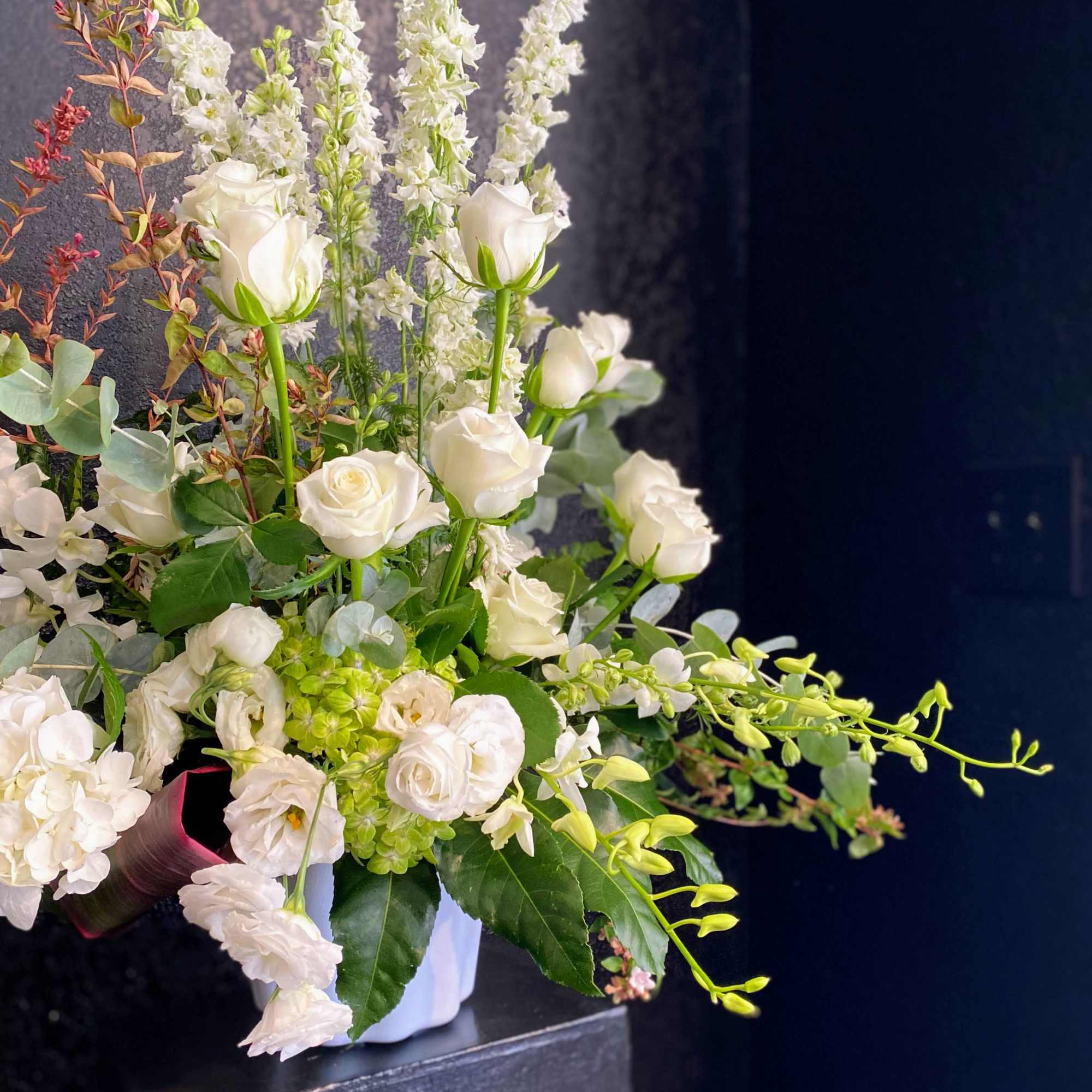 Tall white floral arrangement with roses, hydrangeas, orchids, and stock in a white vase