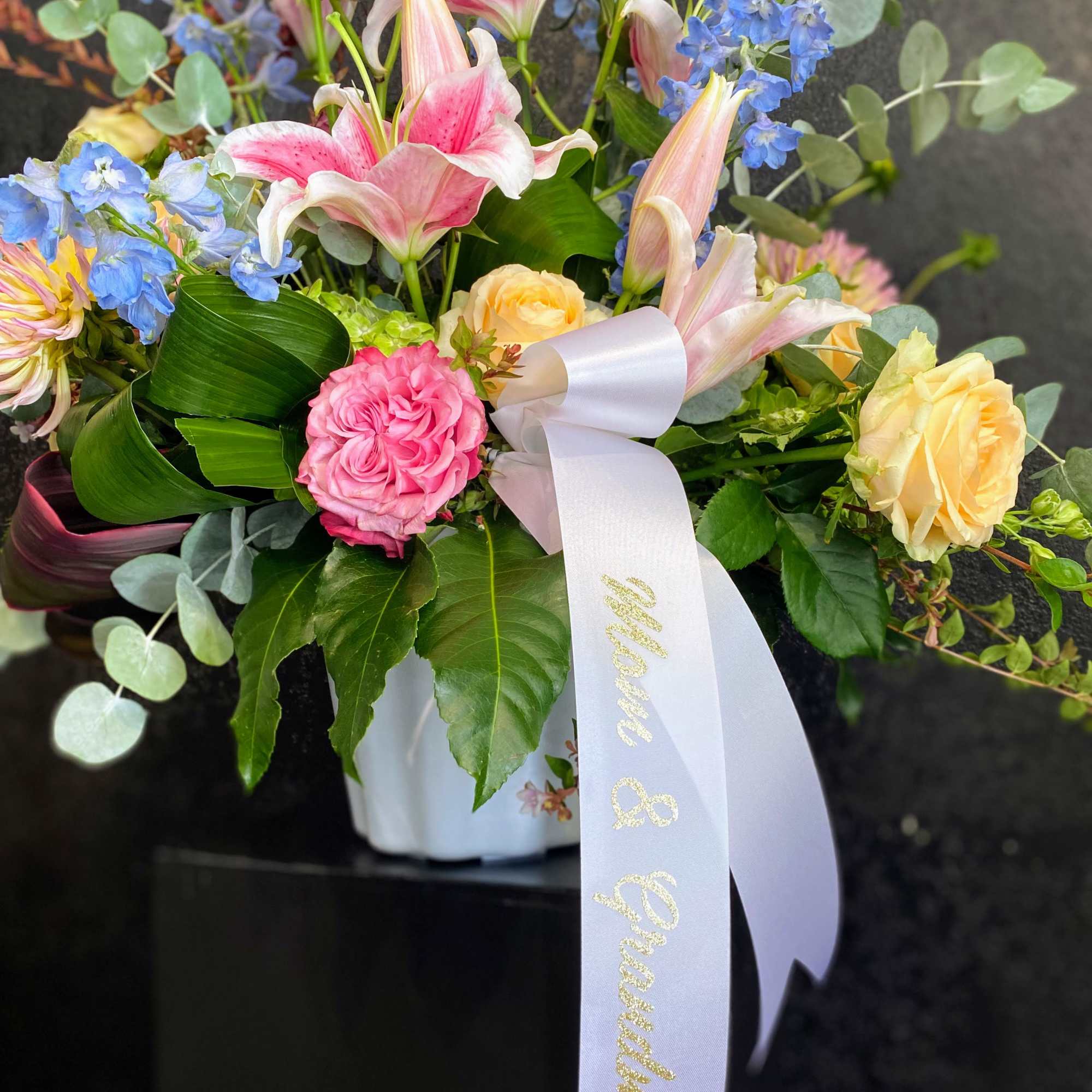 Pink lilies, roses, and blue delphinium in a white vase with a ribbon reading “Mom & Grandma”.