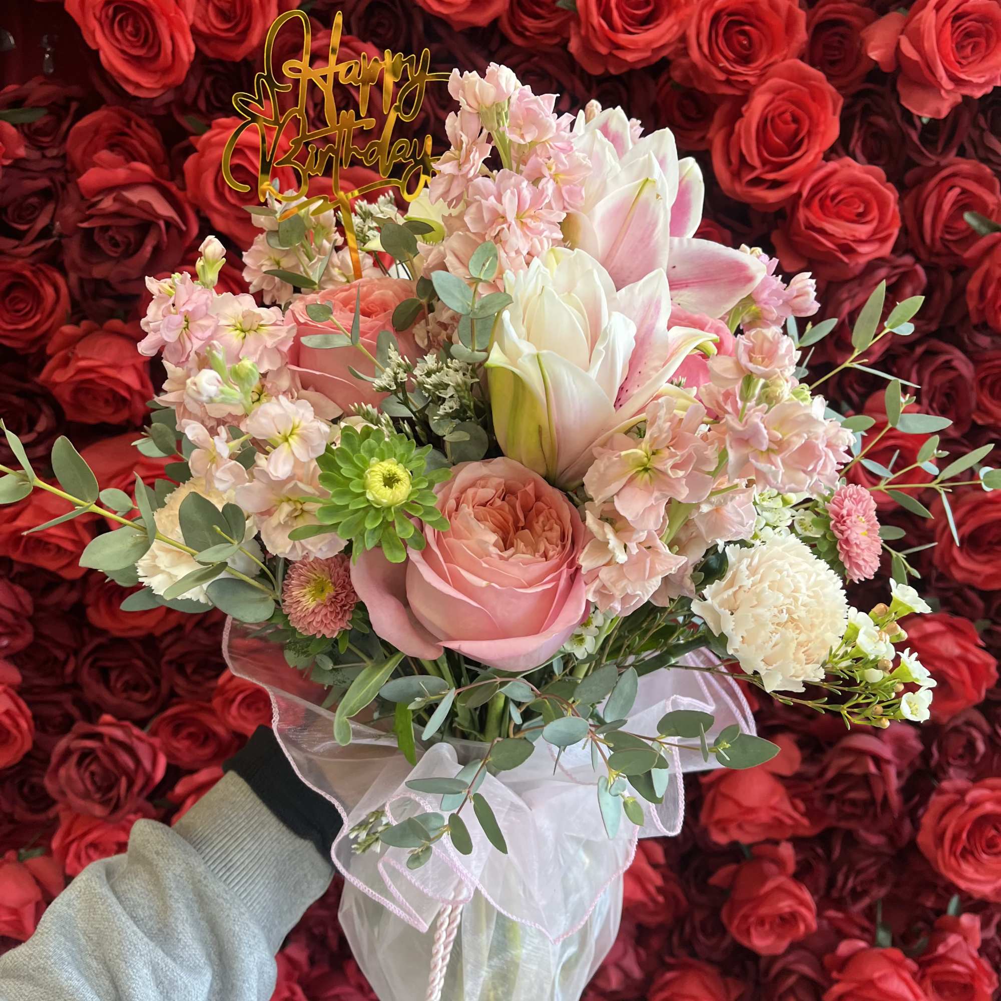 Pink and white bouquet with a birthday topper against a wall of red roses