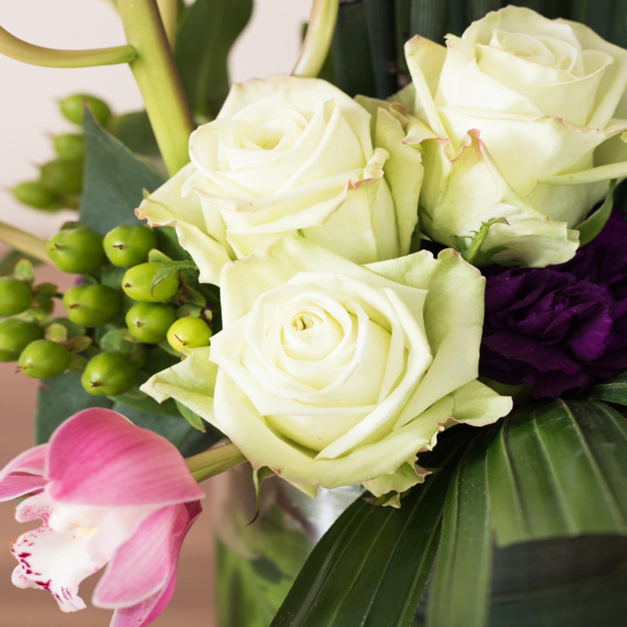 Close-up of an arrangement with pale roses, a pink orchid, green berries, and a dark purple flower in a glass vase