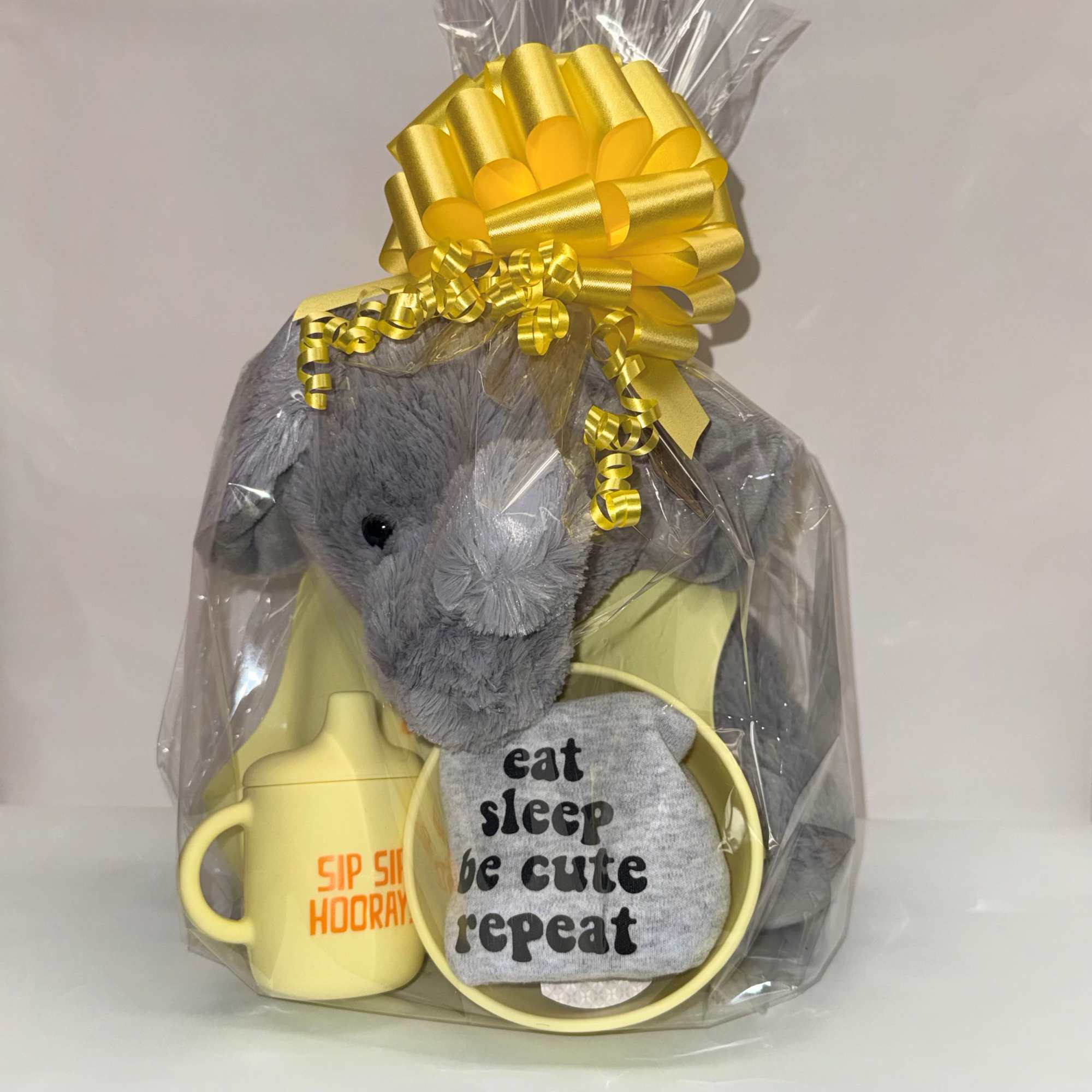 Baby gift set with gray plush elephant, yellow cup, and clothing wrapped in cellophane with a yellow bow