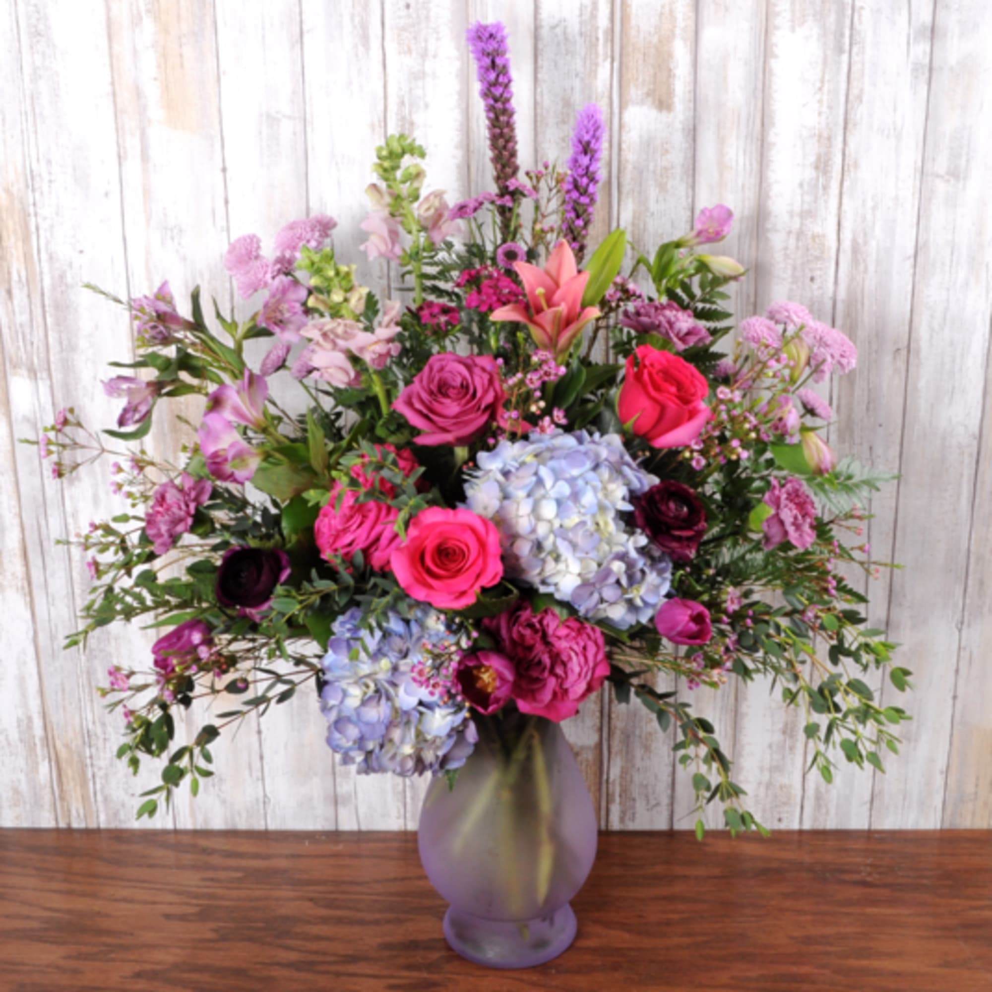 Tall mixed arrangement of pink roses, purple blooms, and blue hydrangeas in a frosted purple vase